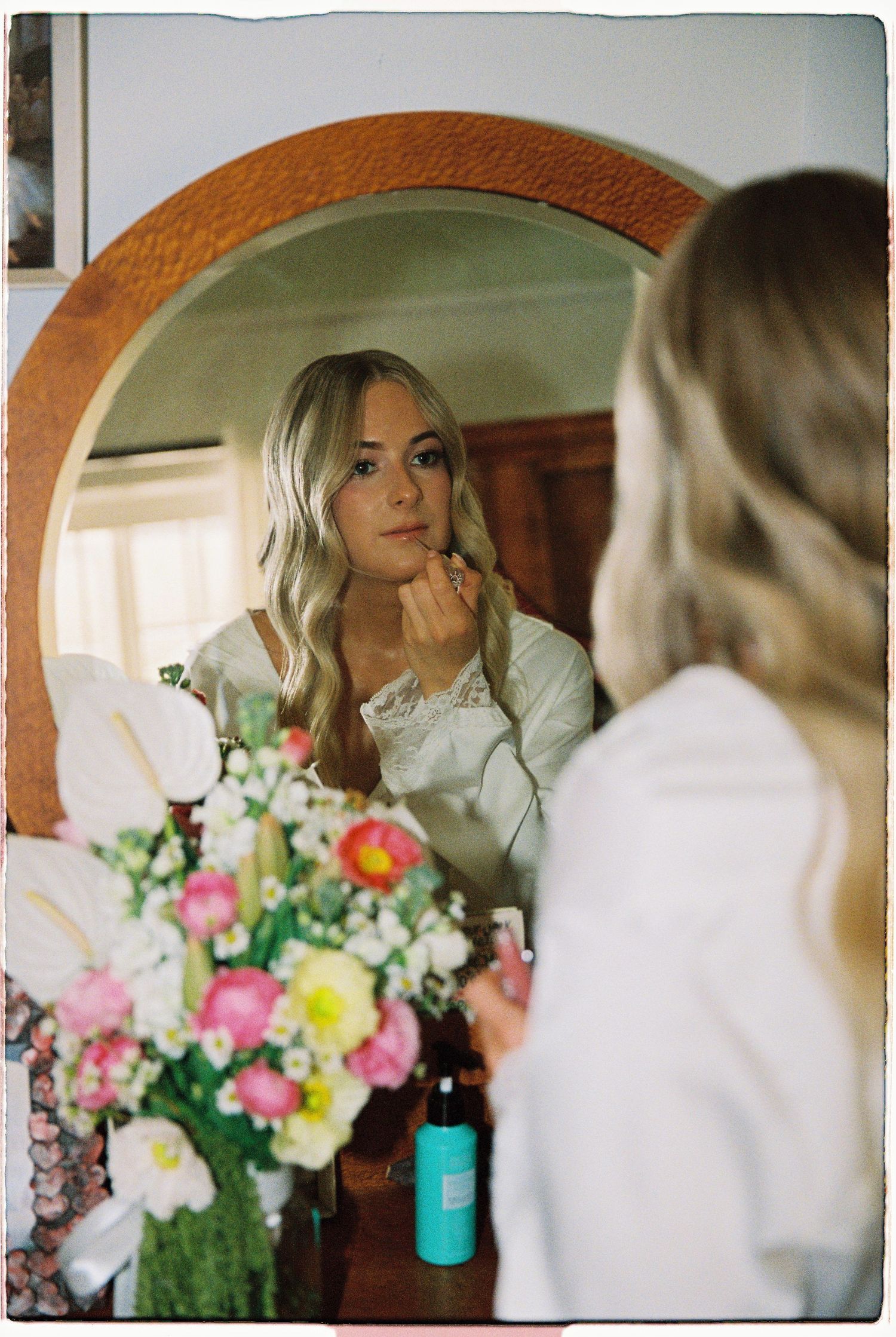 Blonde bride in white robe applying lipstick in round mirror with flowers below.