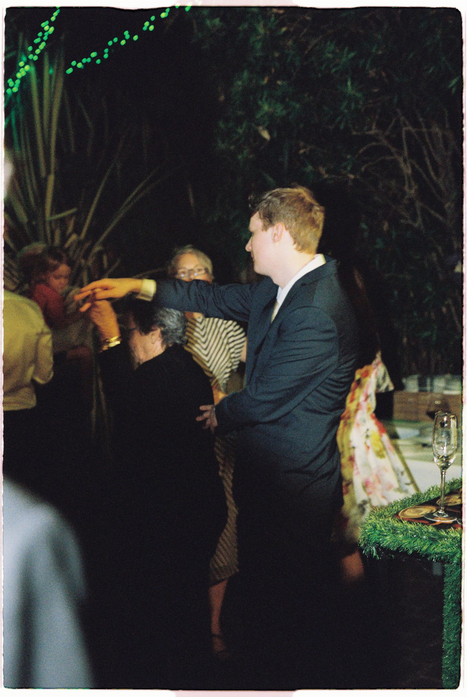 Guests dance and celebrate at an outdoor evening wedding reception surrounded by lush greenery and string lights.