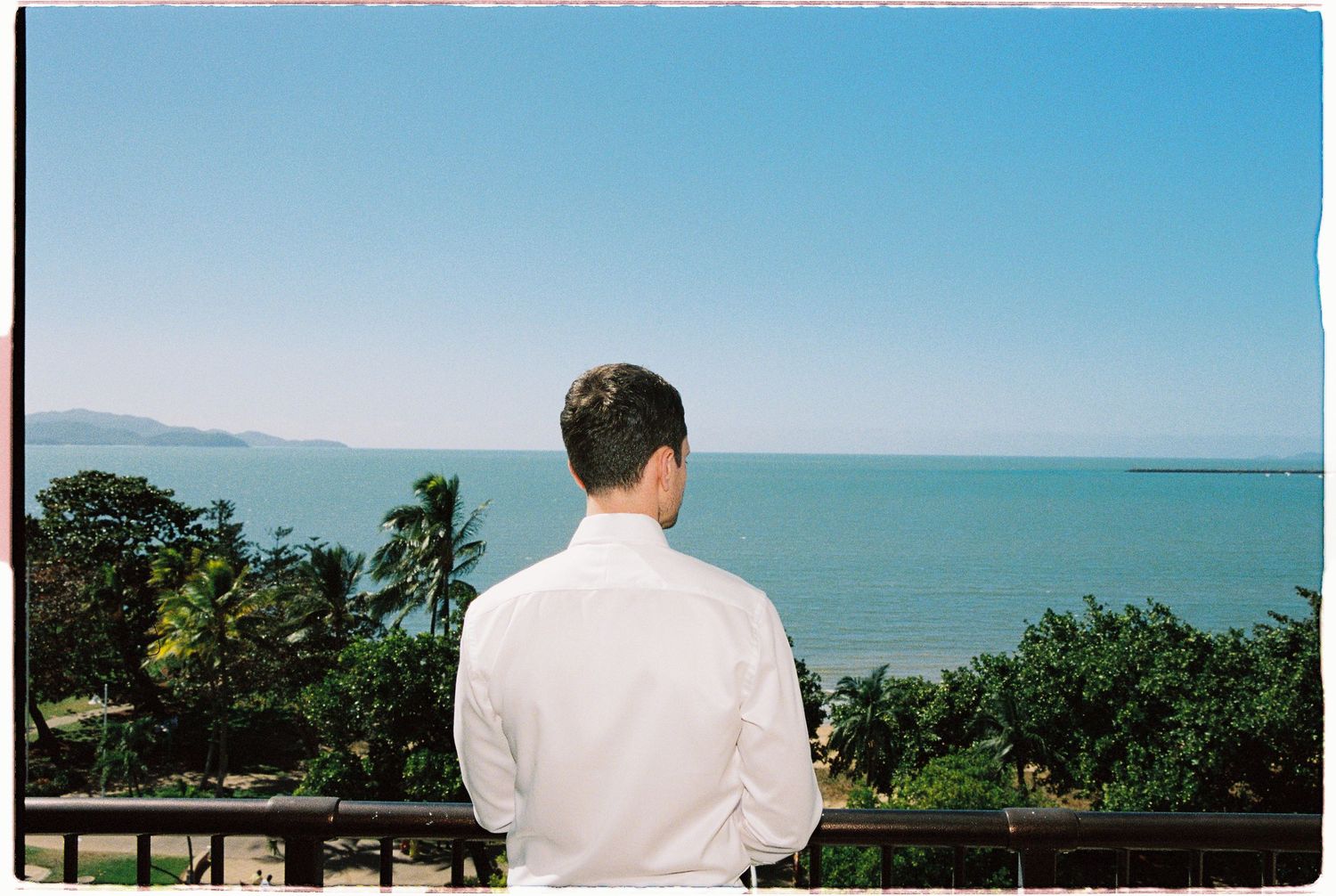 Man in white shirt stands on balcony overlooking tropical ocean coastline with lush green trees under clear blue sky.