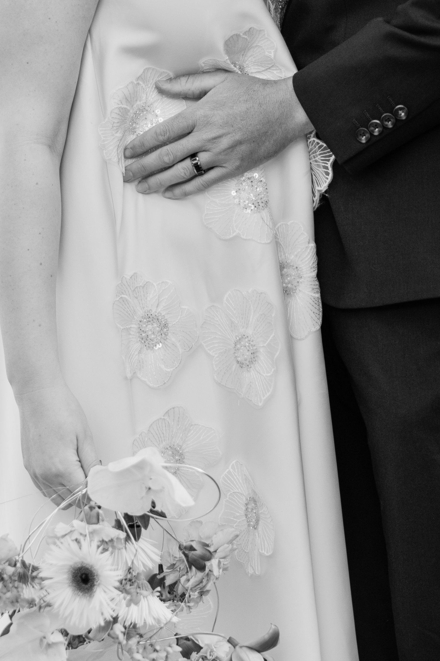 Black and white wedding photo of bride in floral gown holding bouquet, groom's hand with ring resting on her waist.