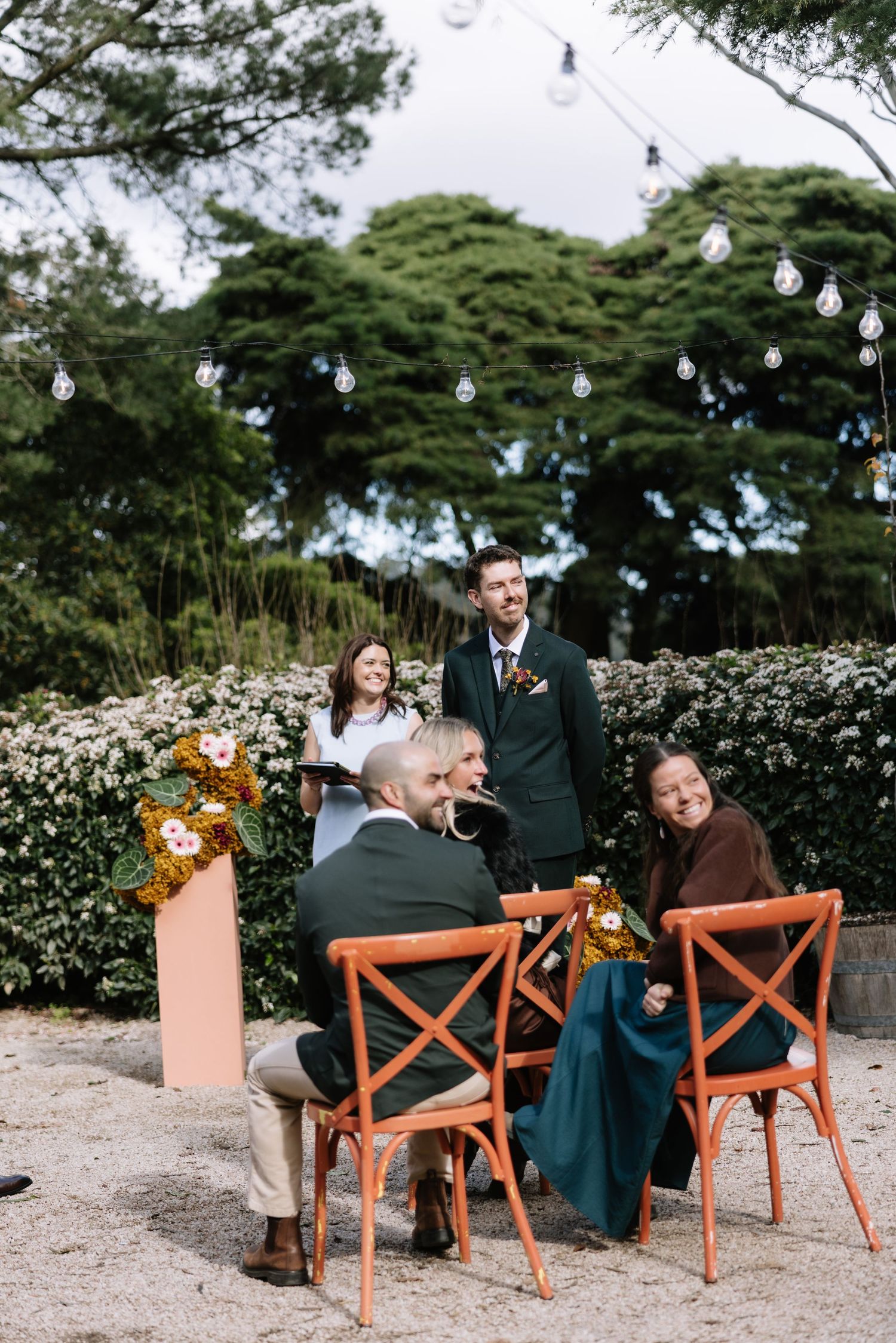 Outdoor wedding ceremony with couple signing documents at orange chairs, officiant standing, string lights and greenery backdrop.