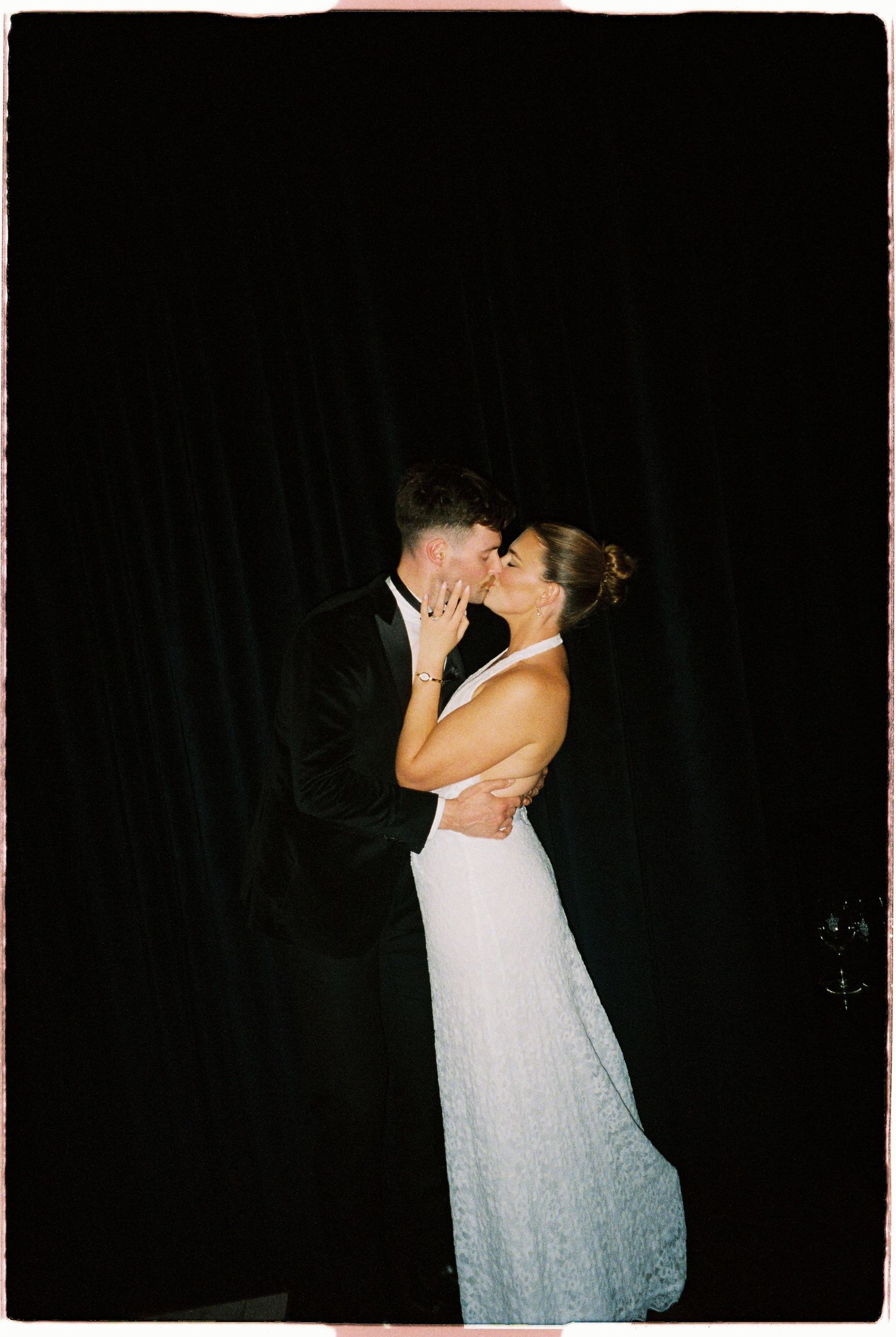 Bride and groom sharing romantic first kiss in wedding attire against dramatic black curtain backdrop.