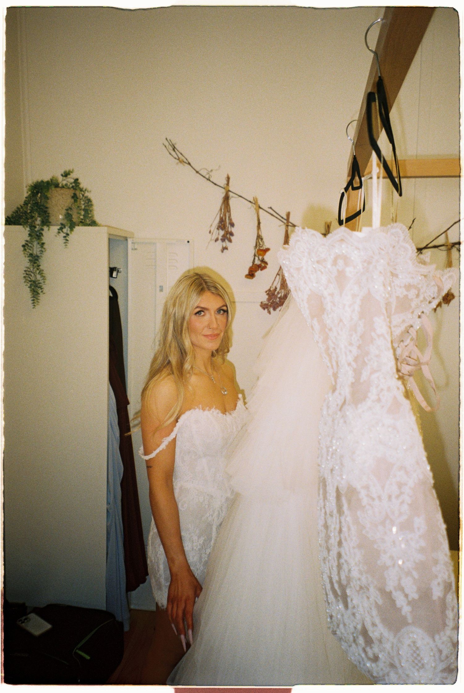 Blonde woman in a white lace wedding dress stands beside a hanging bridal gown in a boutique fitting room.