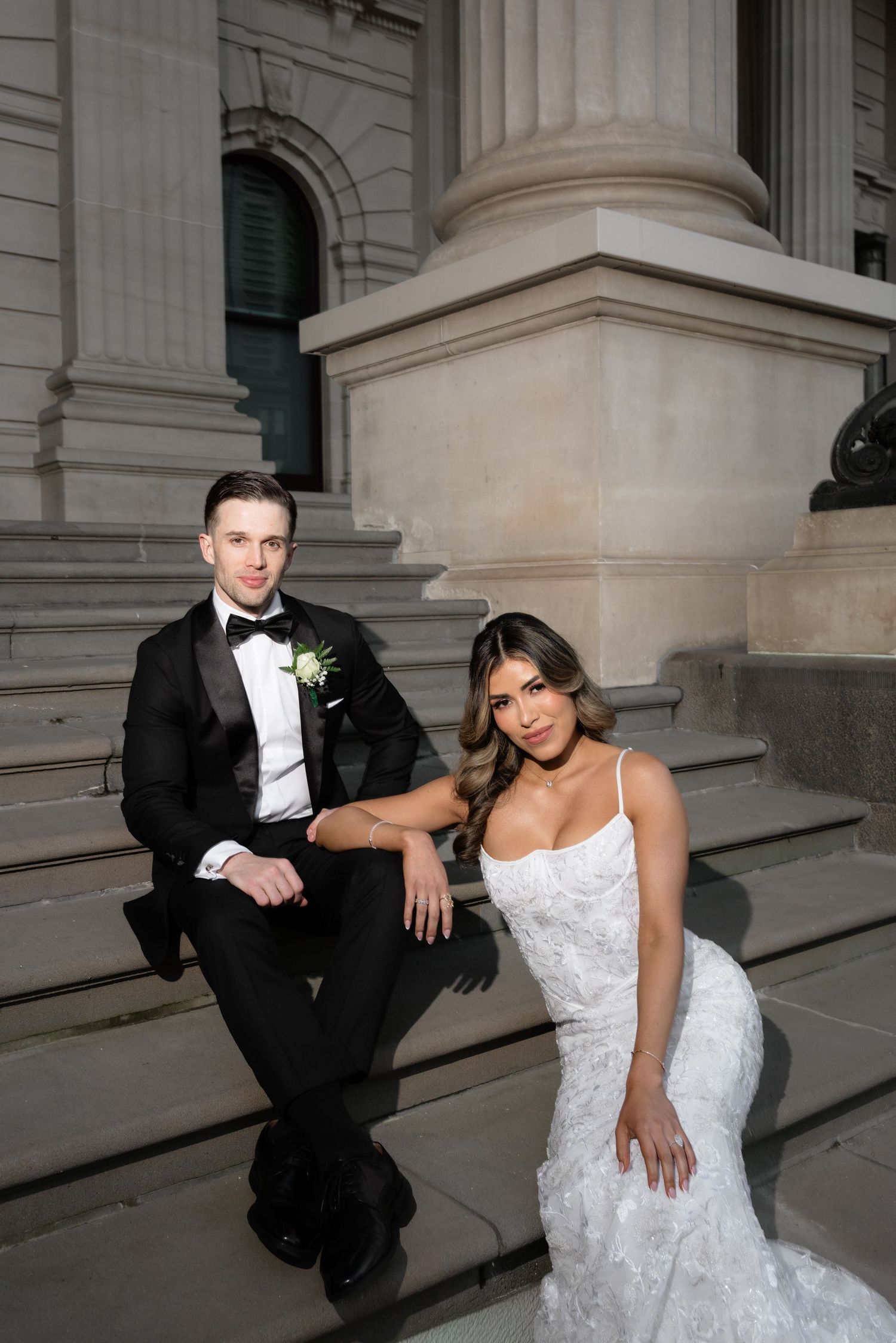 Wedding couple seated on steps by stone column, bride in fitted lace gown, groom in black bow tie tuxedo.