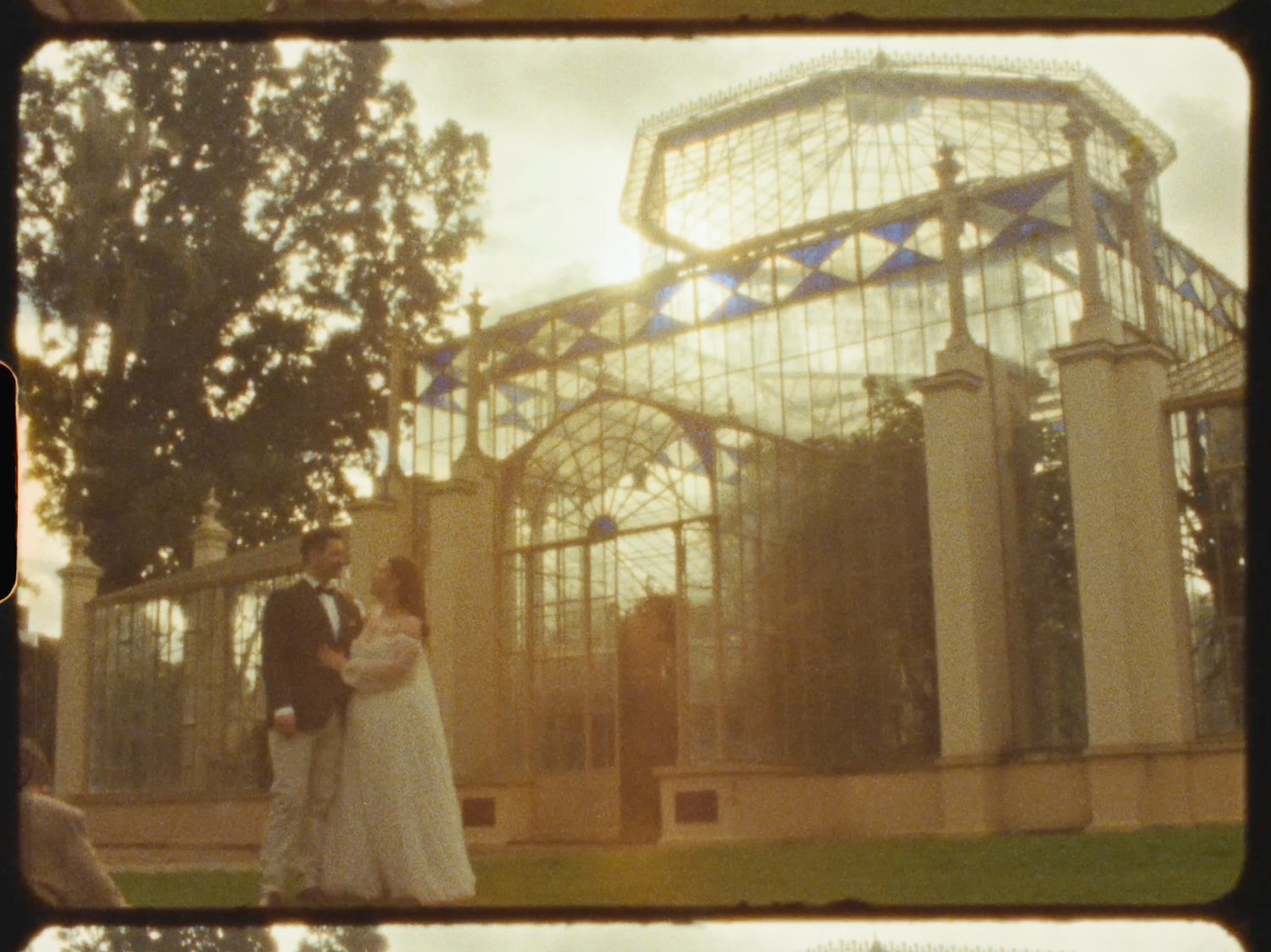 A bride and groom embrace before an ornate Victorian glass greenhouse in a golden-lit garden setting.