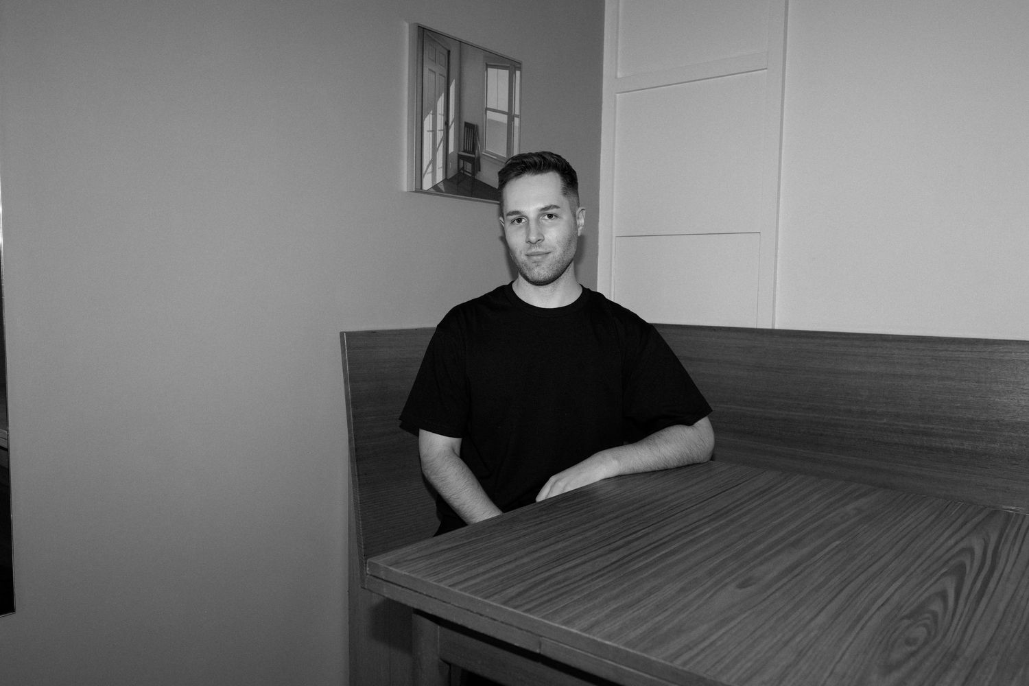 Black and white photo of a man in a black t-shirt sitting at a wooden table in a modern interior.