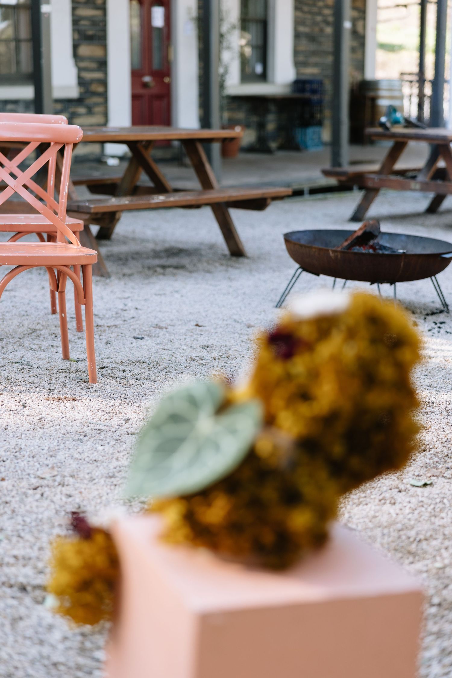 Outdoor patio with pink chairs, wooden picnic tables, a fire pit, and yellow floral arrangement on a pink pedestal.