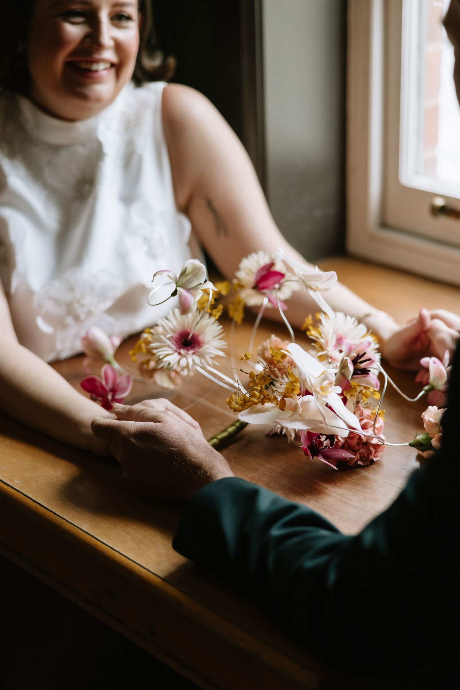 Smiling bride in white dress holding colorful pink and gold floral bouquet at wooden table near window.