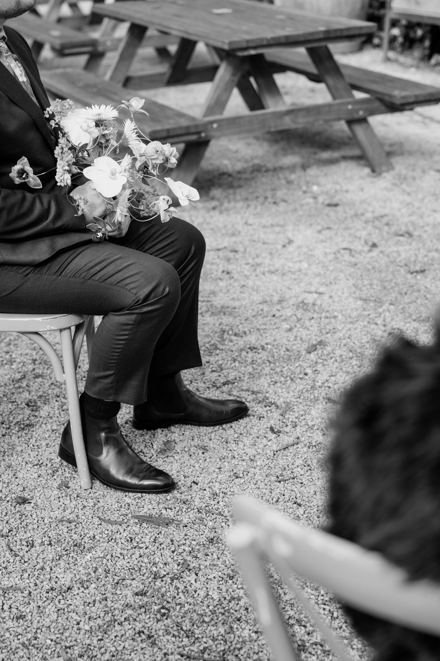 Black and white photo of a seated person in a suit holding a wedding bouquet, with picnic tables visible in the background.