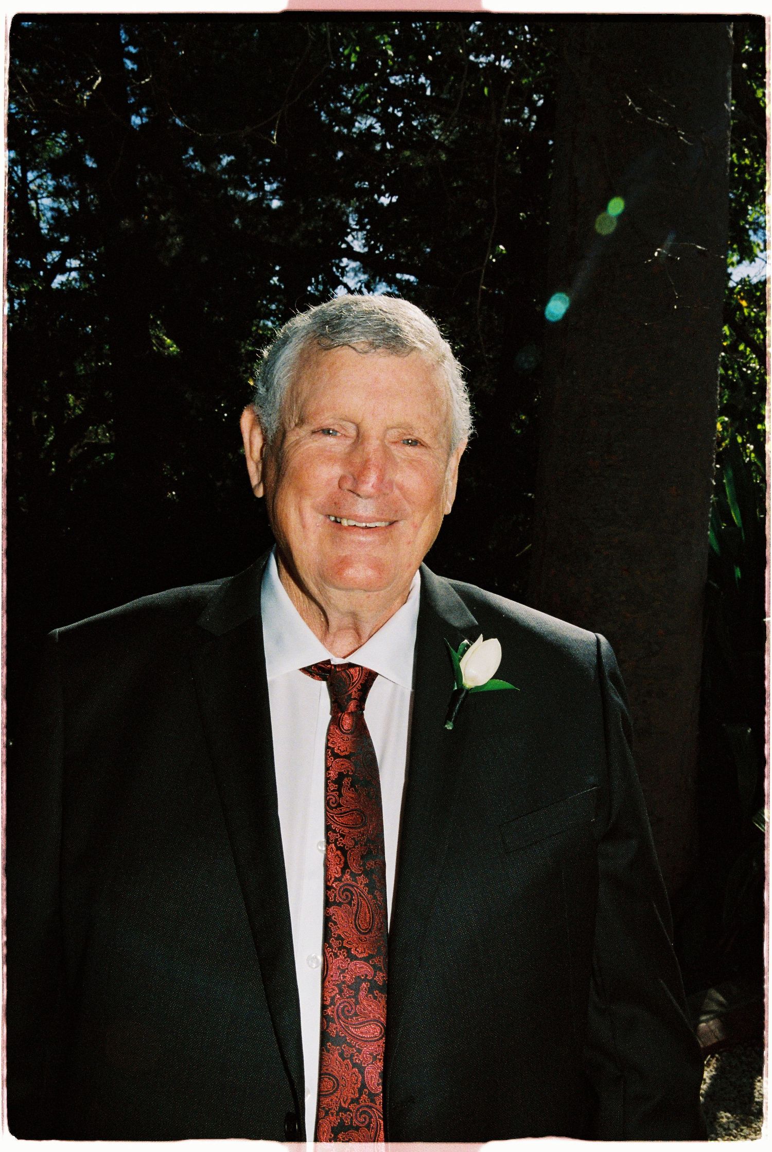 Elderly man in black suit with red floral tie and white boutonniere smiling outdoors among trees.