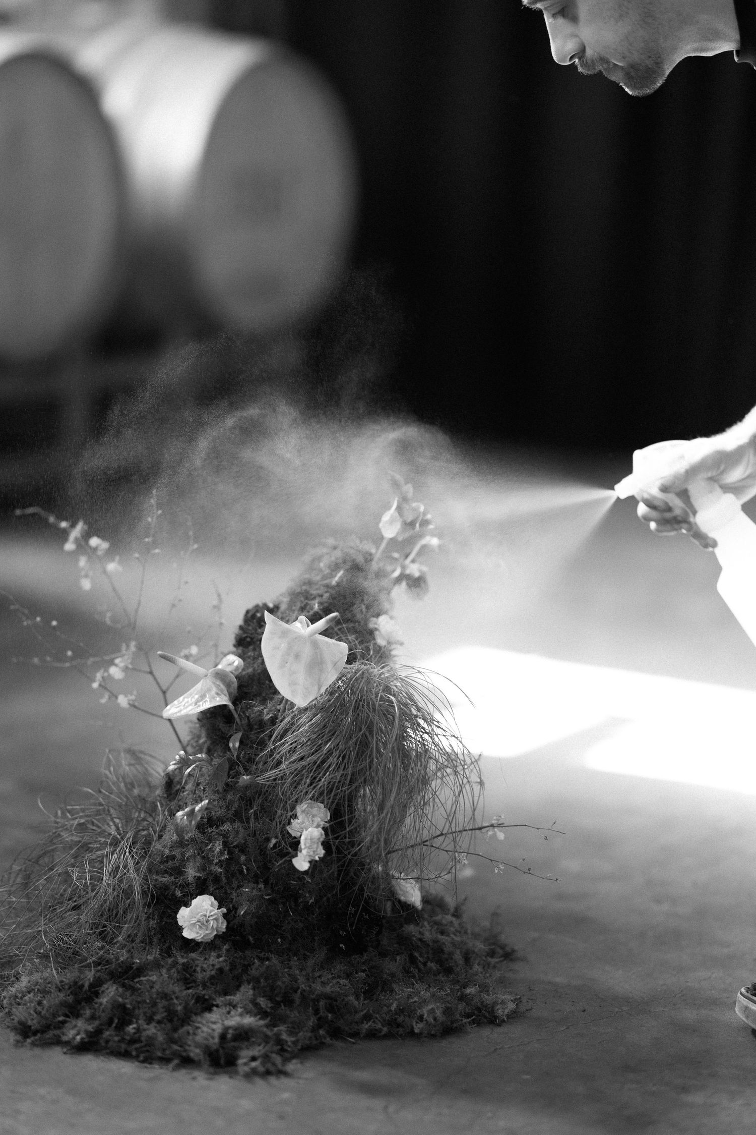 Black and white photo of florist misting moss and anthurium floral arrangement with wine barrels in background.
