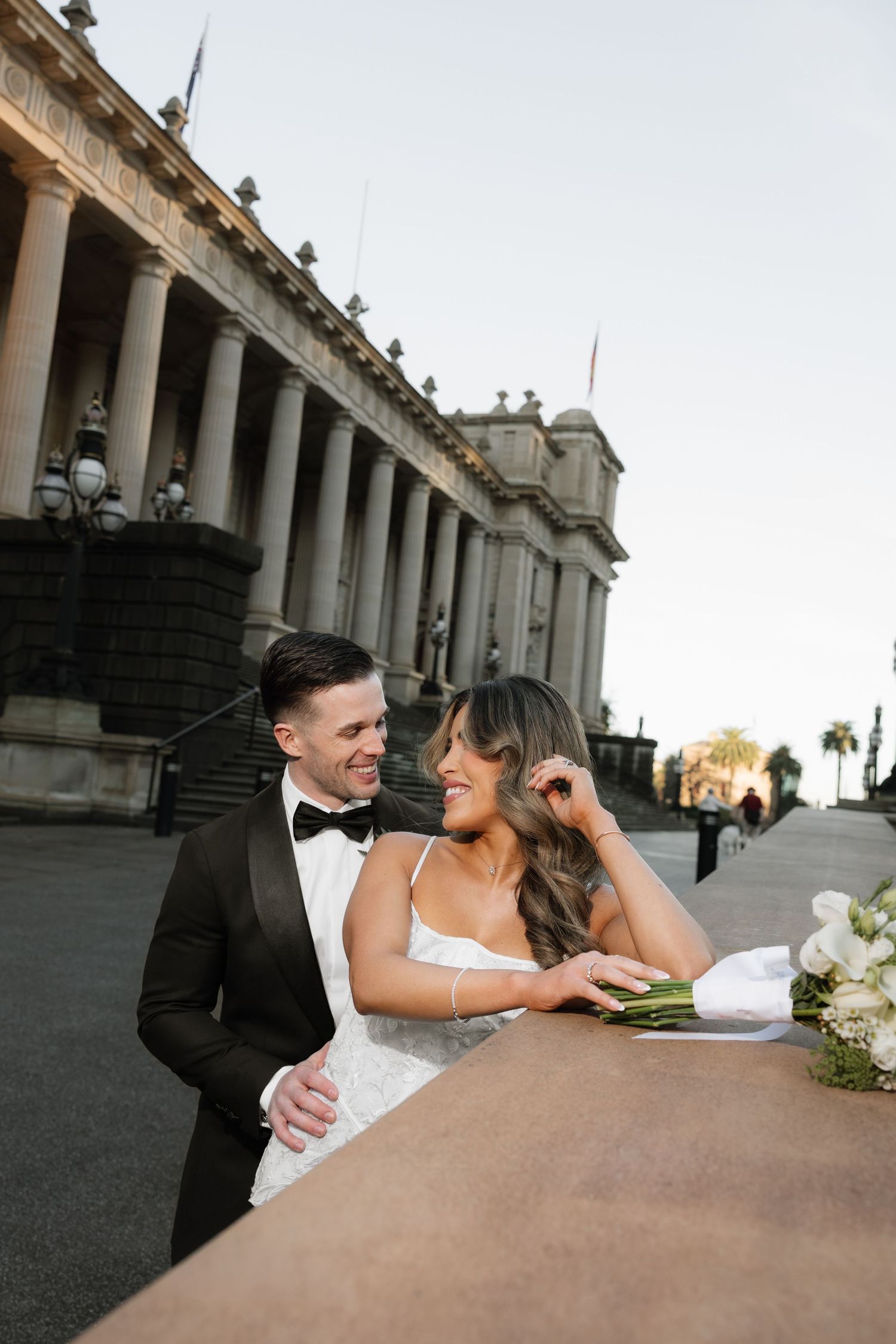 Bride and groom share a romantic moment outside a grand columned building, she in a white gown, he in a black tuxedo.