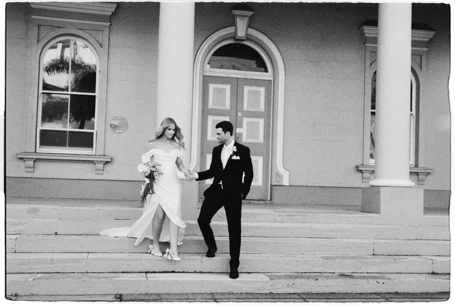 Black and white photo of bride and groom walking down steps of elegant building with white columns.