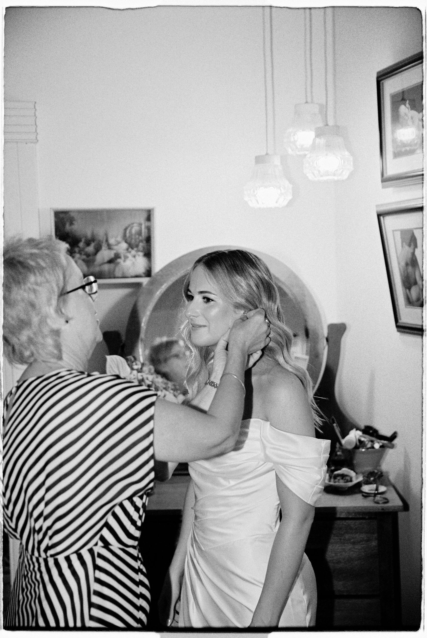 Black and white photo of an older woman helping a bride put on earrings before her wedding ceremony.
