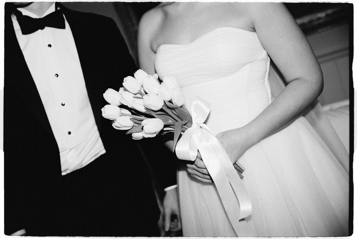 Black and white wedding photo of bride holding white tulip bouquet with ribbon, standing beside groom in tuxedo.