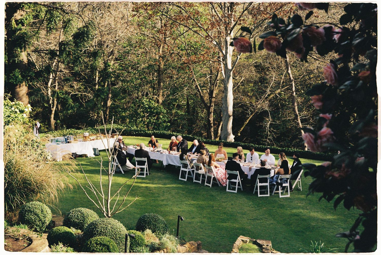 Outdoor garden wedding reception with guests seated at long decorated tables on a green lawn surrounded by trees.