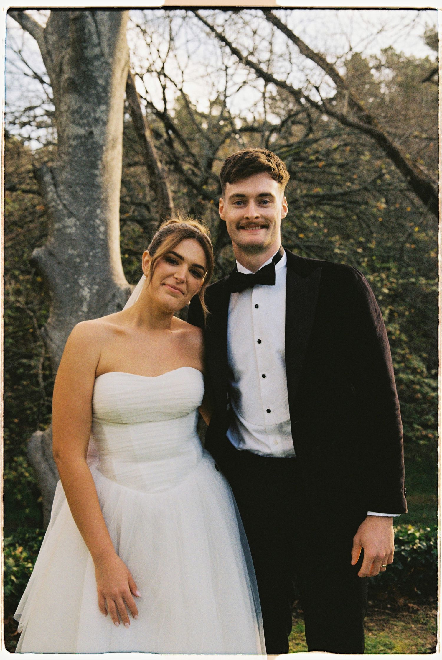 Bride in strapless white gown and groom in black tuxedo posing together outdoors at their wedding.