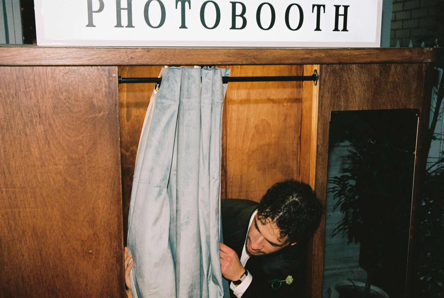 Guest in black suit ducking into vintage wooden photo booth at wedding reception.