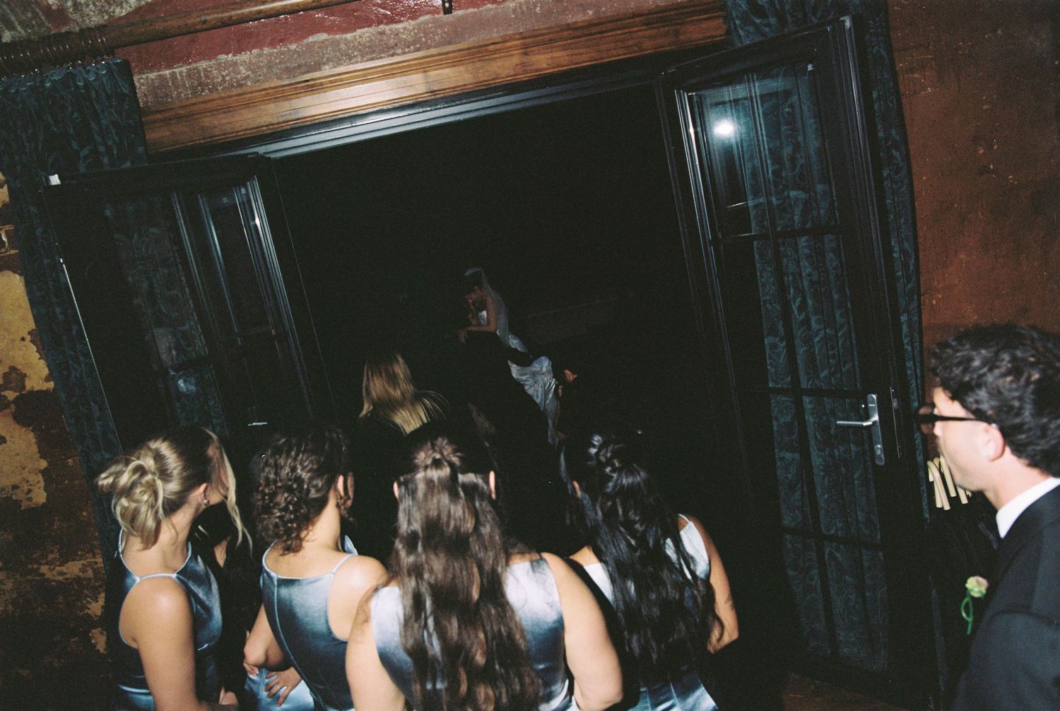 Wedding party in blue bridesmaid dresses gathering at dark ornate doorway entrance.