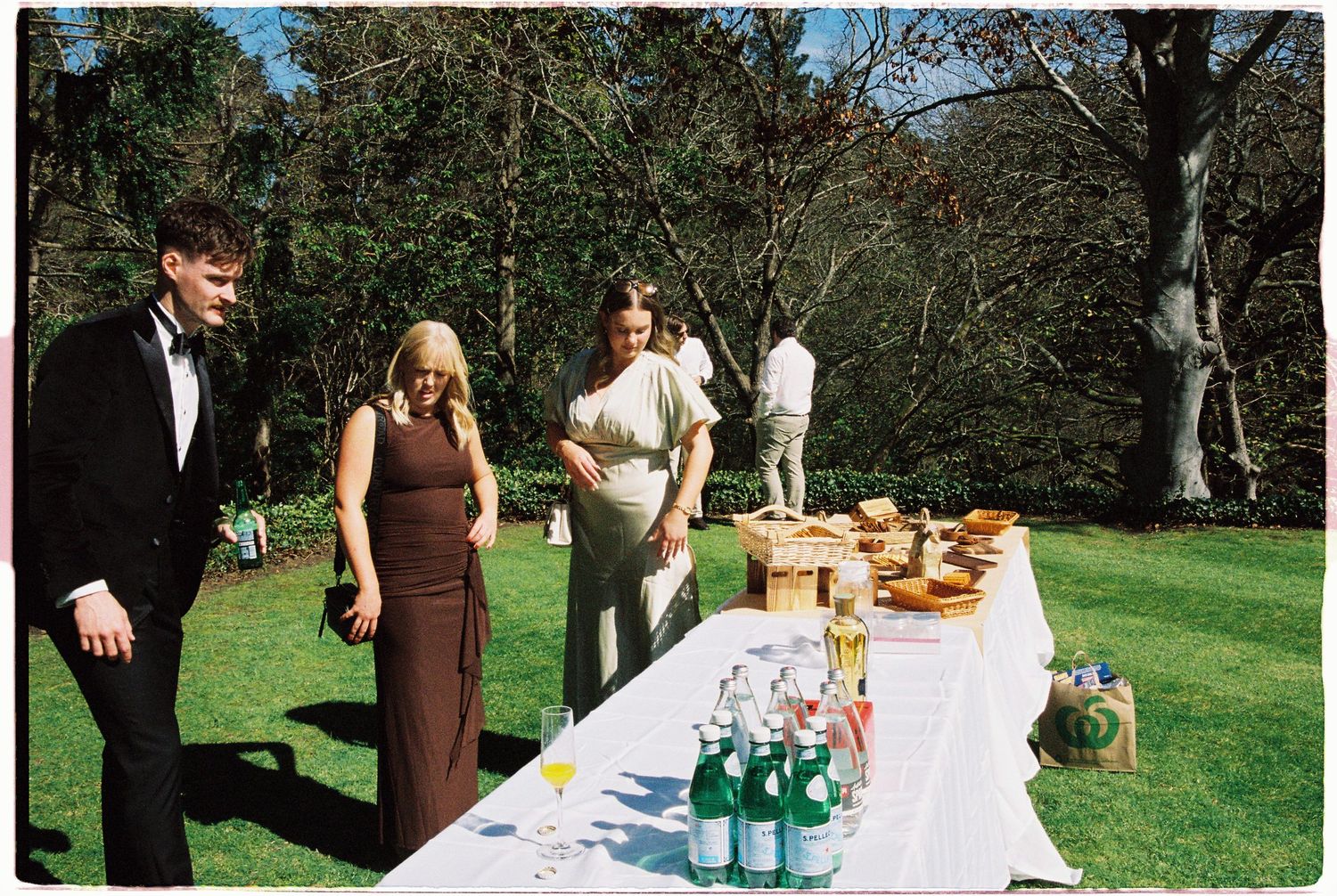 Wedding guests browse food and drinks on a sunlit outdoor reception table in a green garden setting.