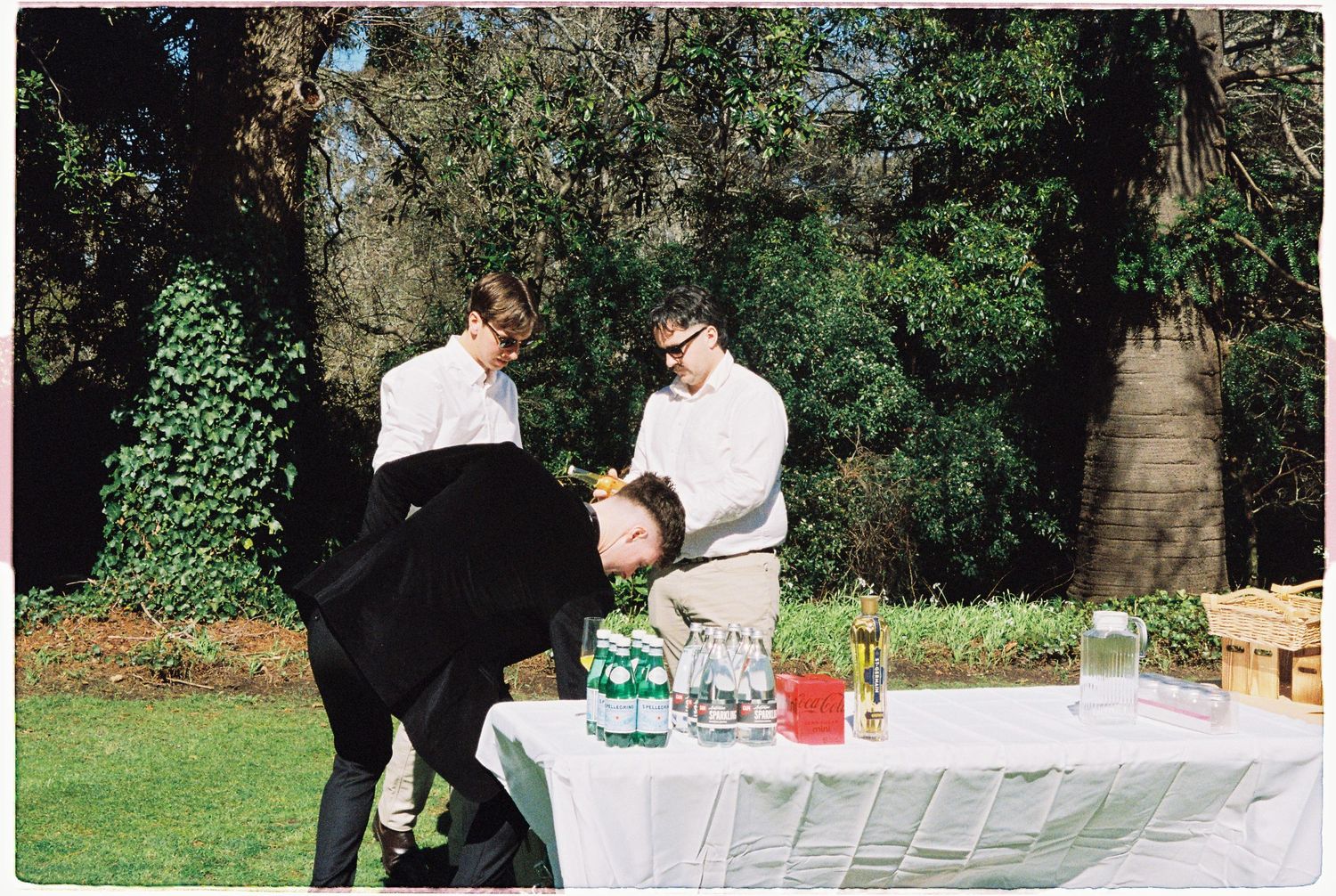Three guests interact at a white-clothed outdoor reception table with drinks and a picnic basket.