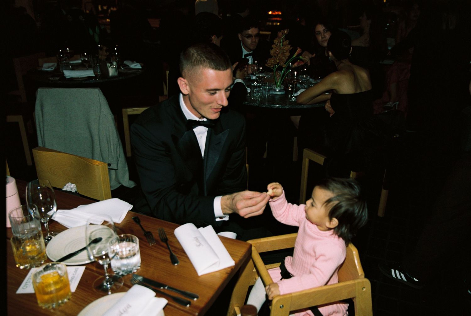 A man in a tuxedo shares a tender moment feeding a baby at a wedding reception.