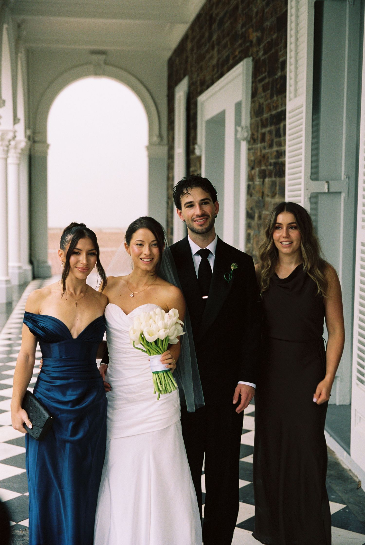 Bride in white gown holding tulips poses with groom in black suit and two bridesmaids on elegant venue colonnade.