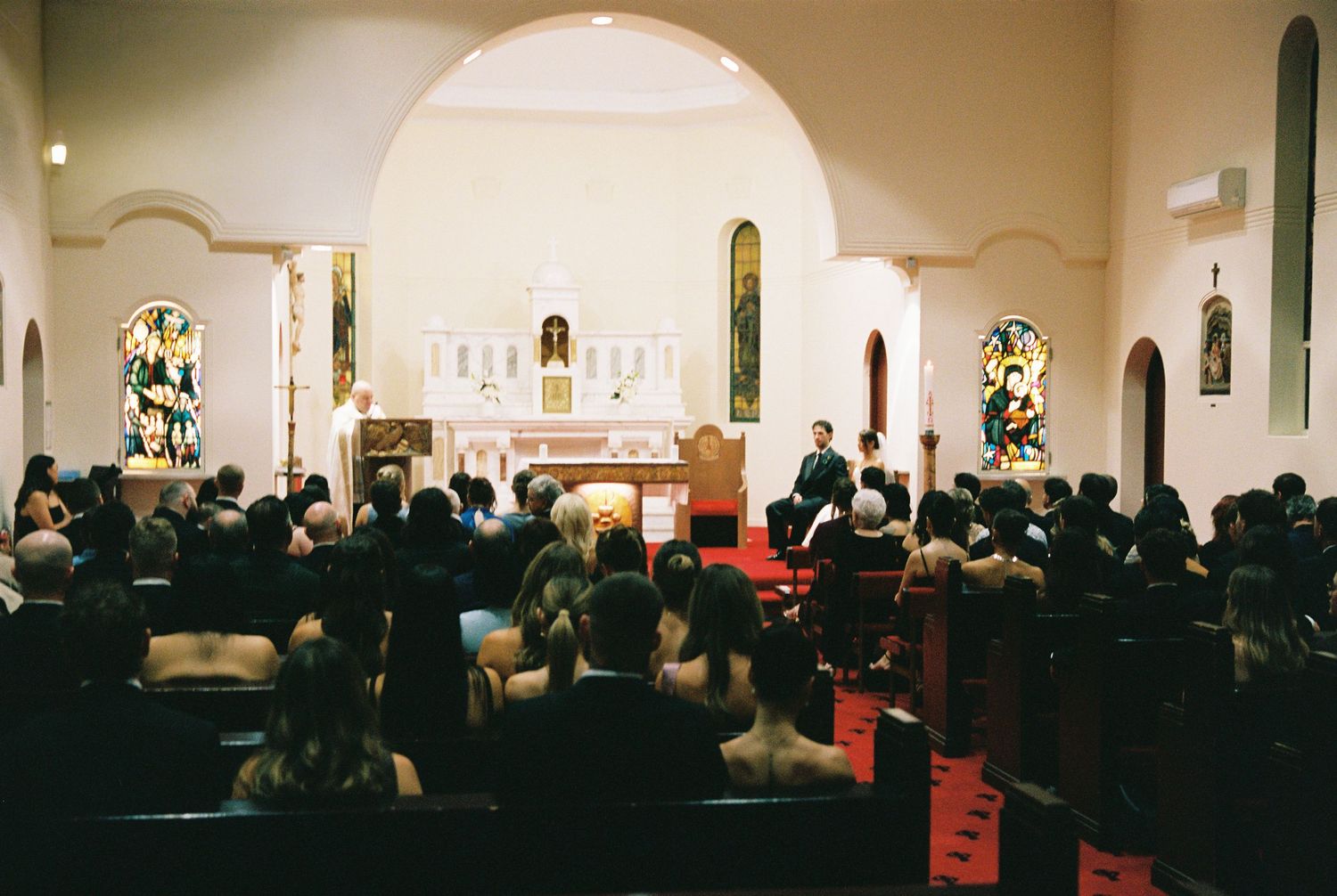 Wedding guests fill church pews during ceremony with colorful stained glass windows.