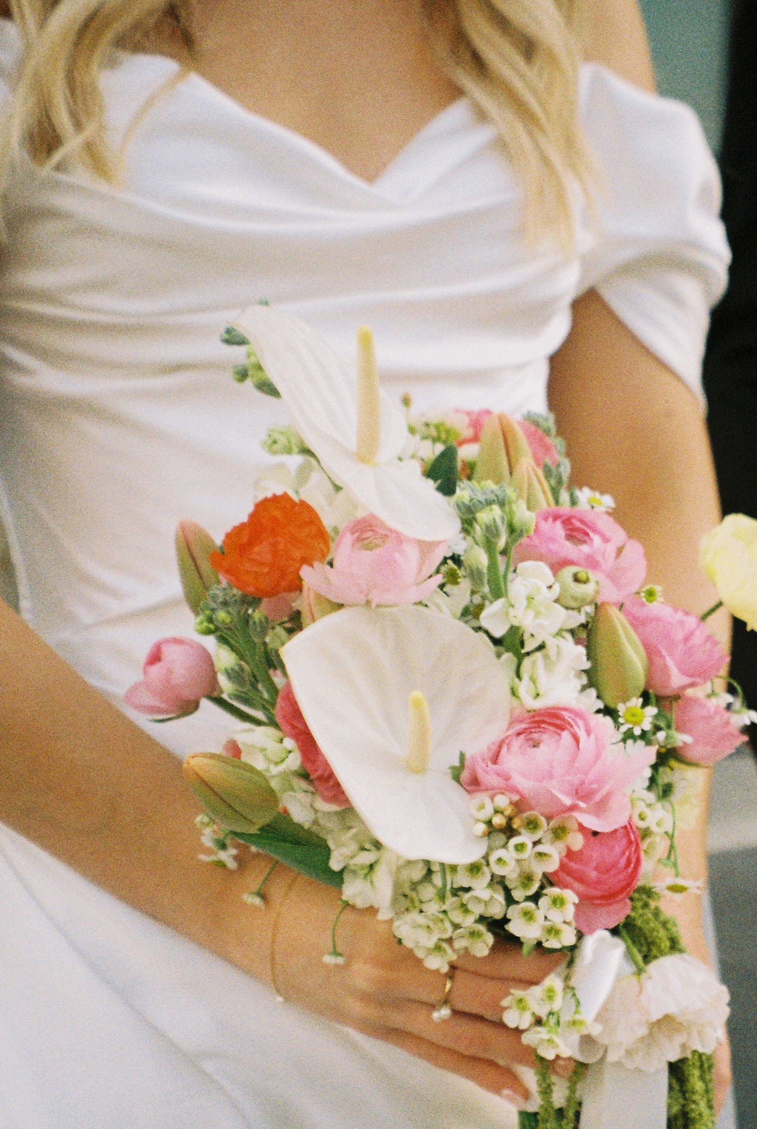 Bride holds a stunning bouquet of pink ranunculus, white anthuriums, and orange flowers in white satin gown.