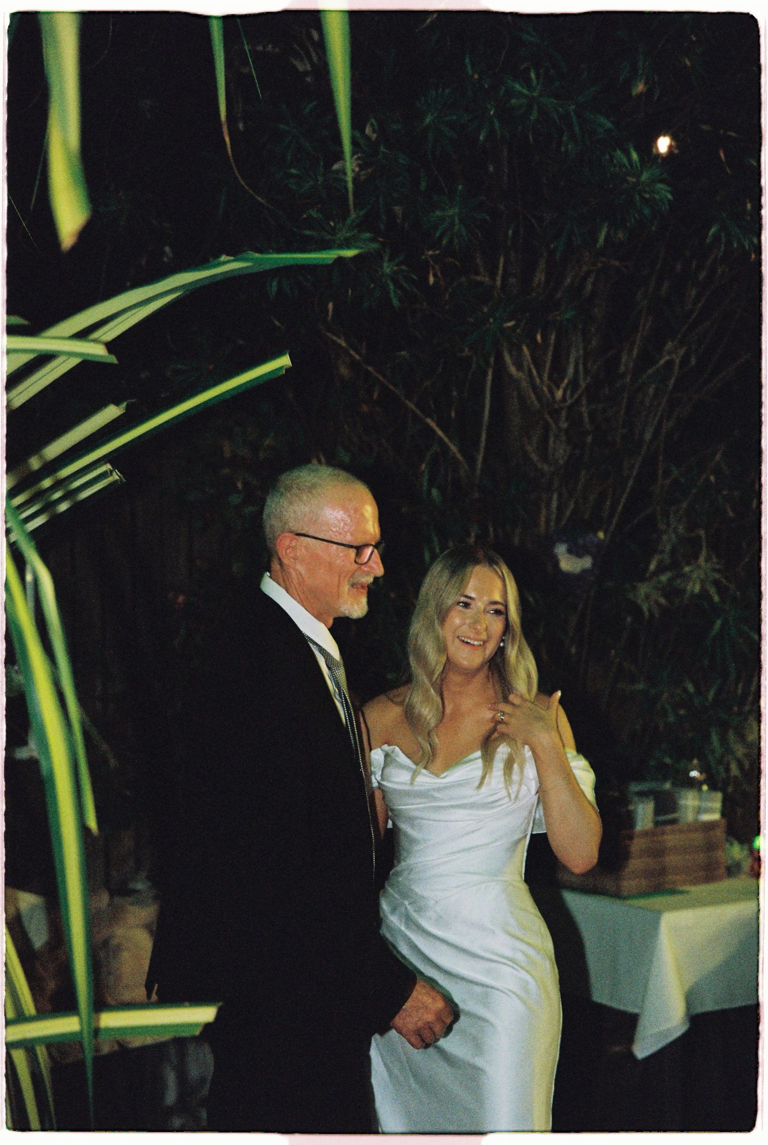 A bride in a white gown smiles while walking with an older man in a suit at an outdoor evening wedding.
