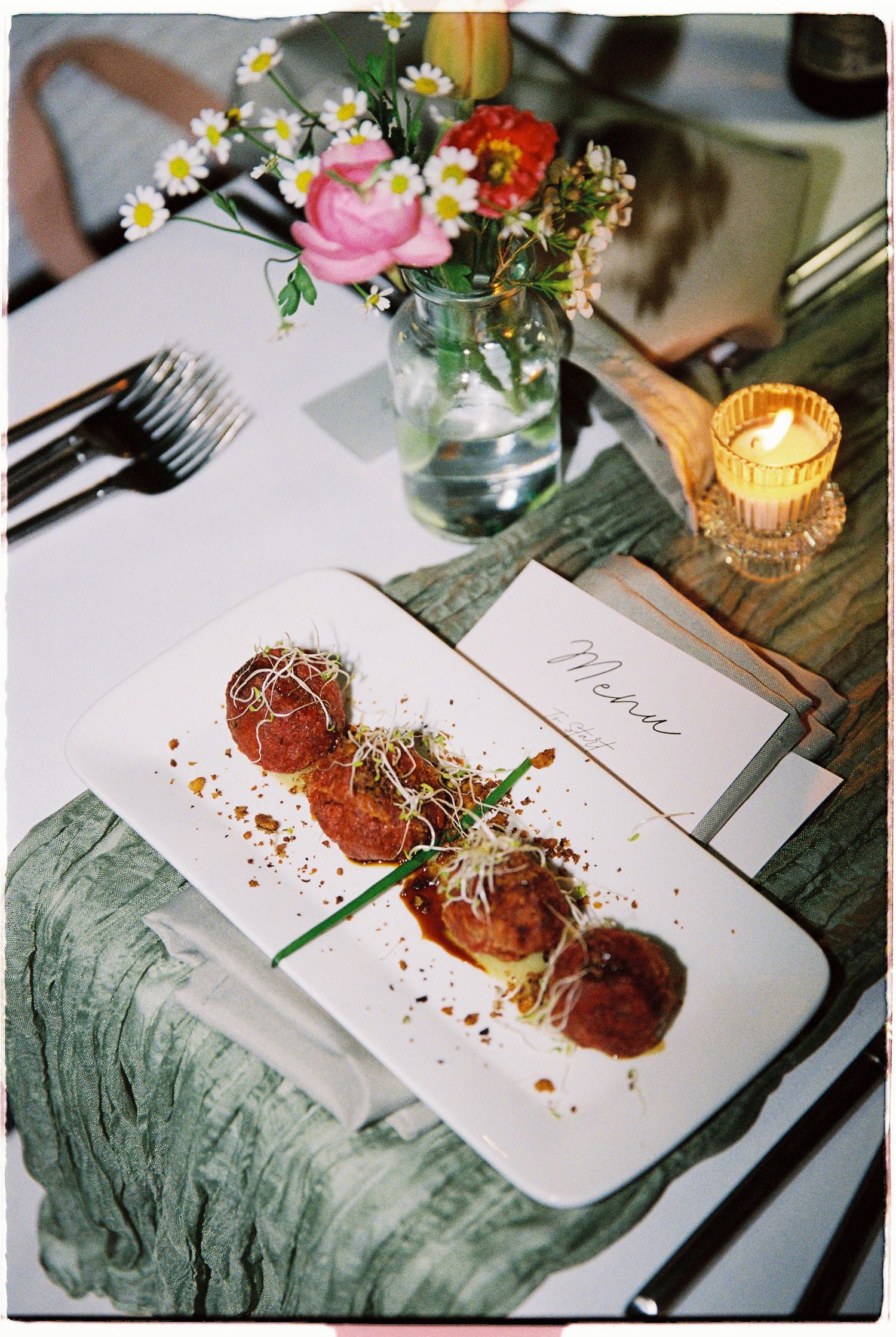 Elegant wedding reception table setting with gourmet appetizers, floral centerpiece, menu card, and glowing candle.