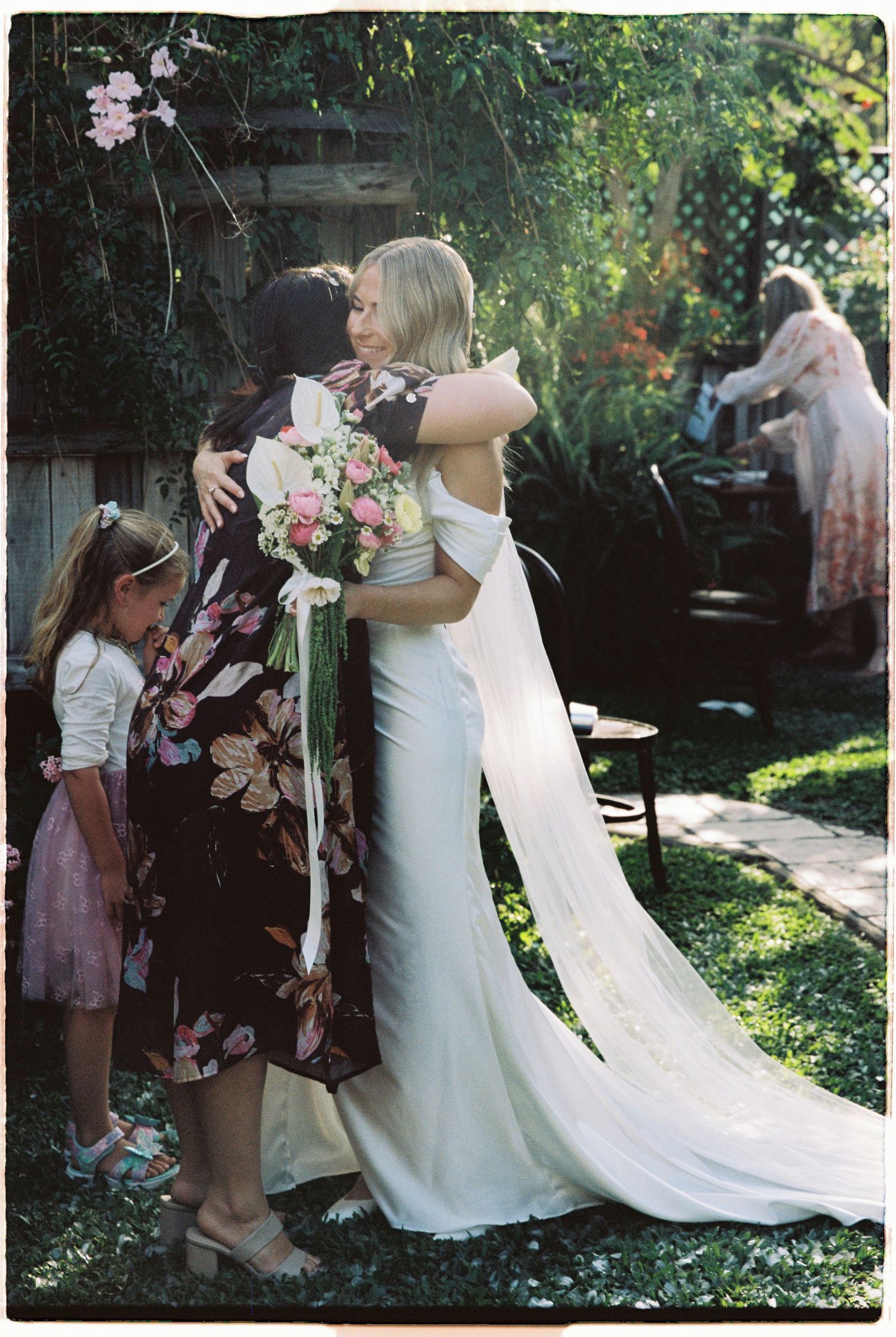 Bride in elegant white gown with long train receives warm hug while holding pink floral bouquet.