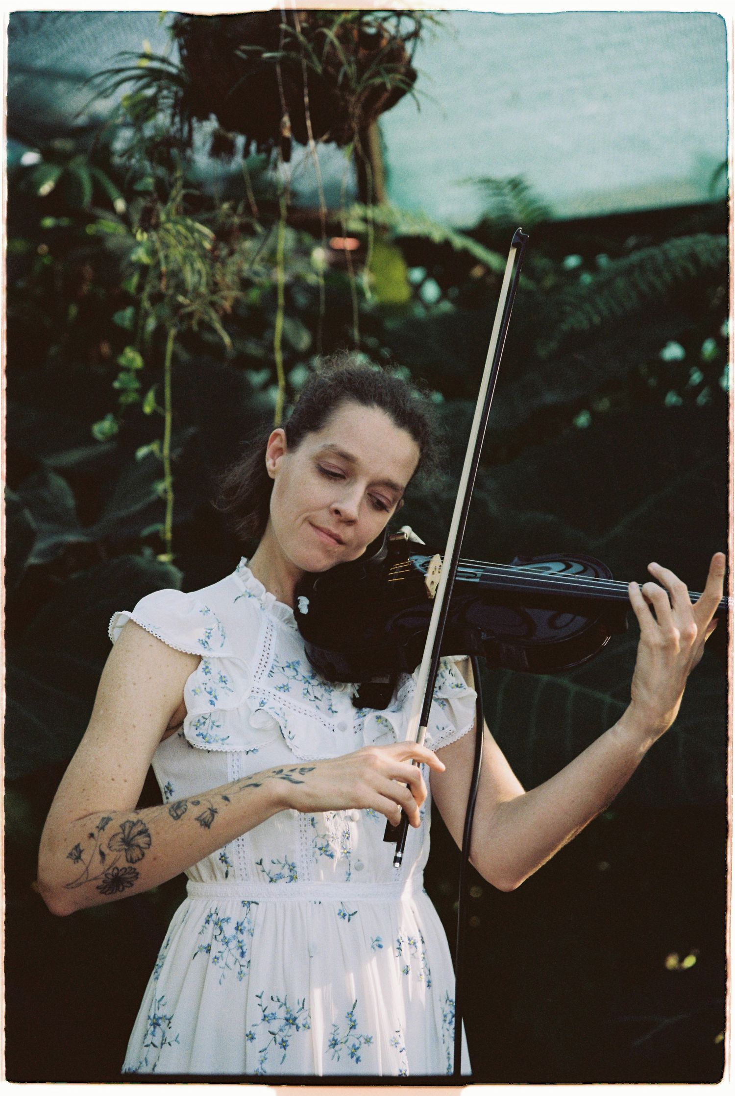 Tattooed woman in floral white dress plays violin surrounded by tropical green plants at wedding ceremony.