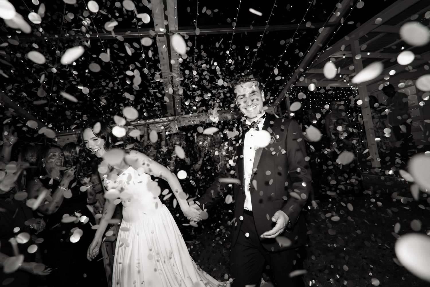 Black and white photo of a bride and groom walking through a shower of white confetti petals.
