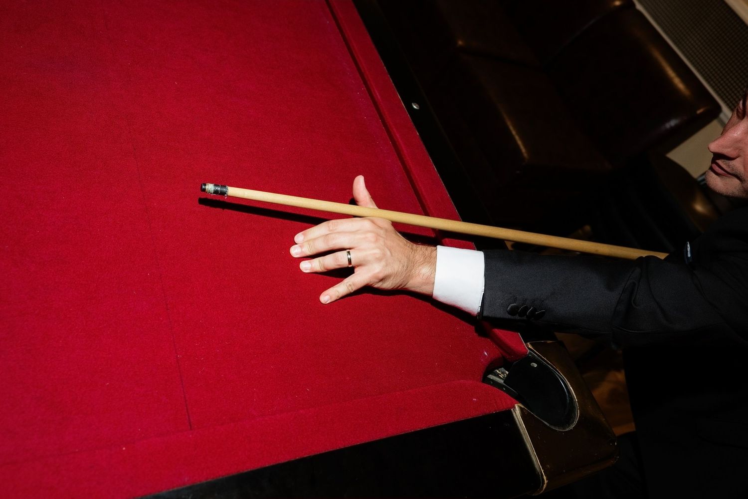 Close-up of a hand holding a pool cue over a red billiard table, wearing a suit and ring.