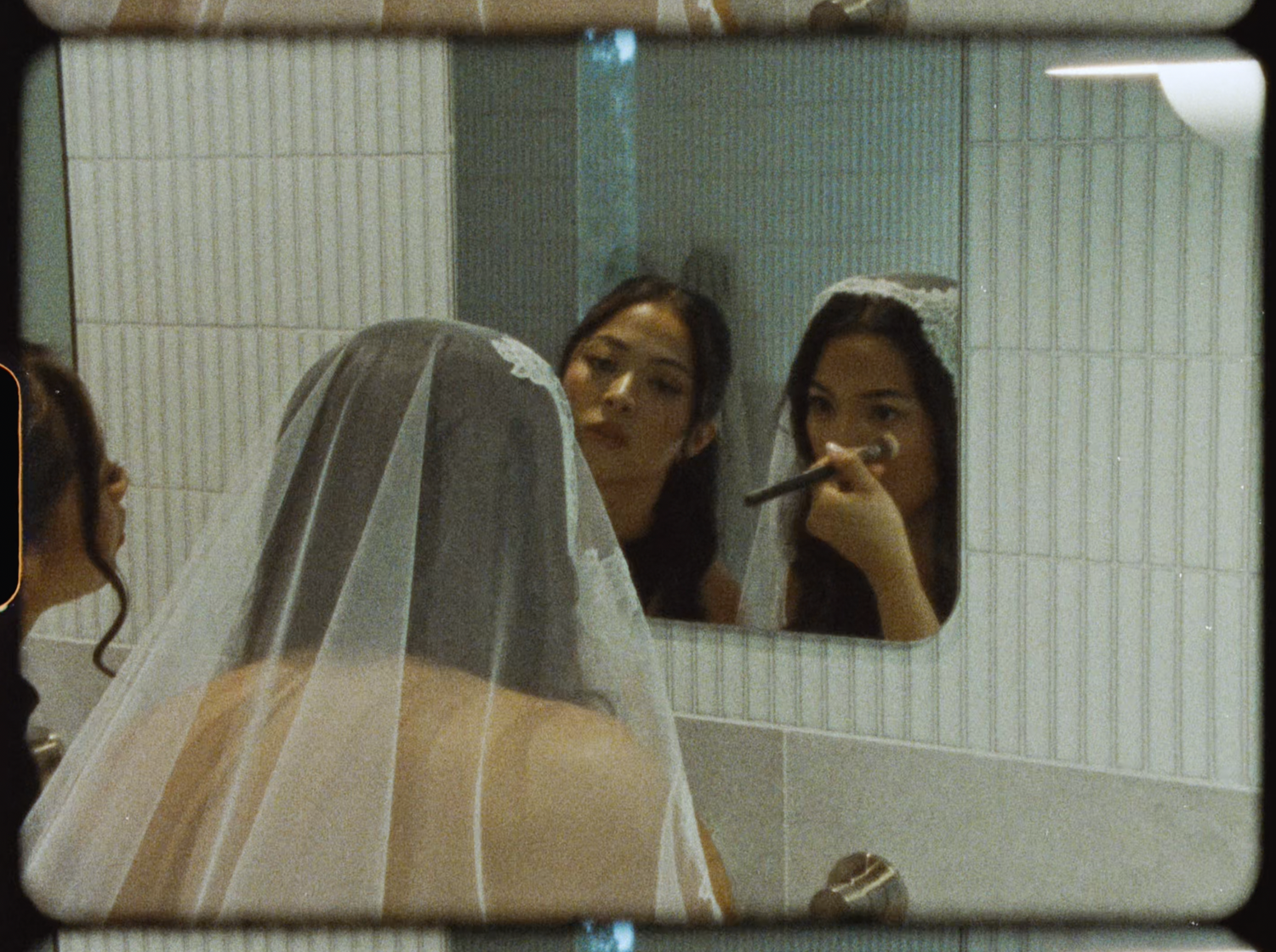 Bride wearing a lace veil gets makeup applied while looking in a bathroom mirror.
