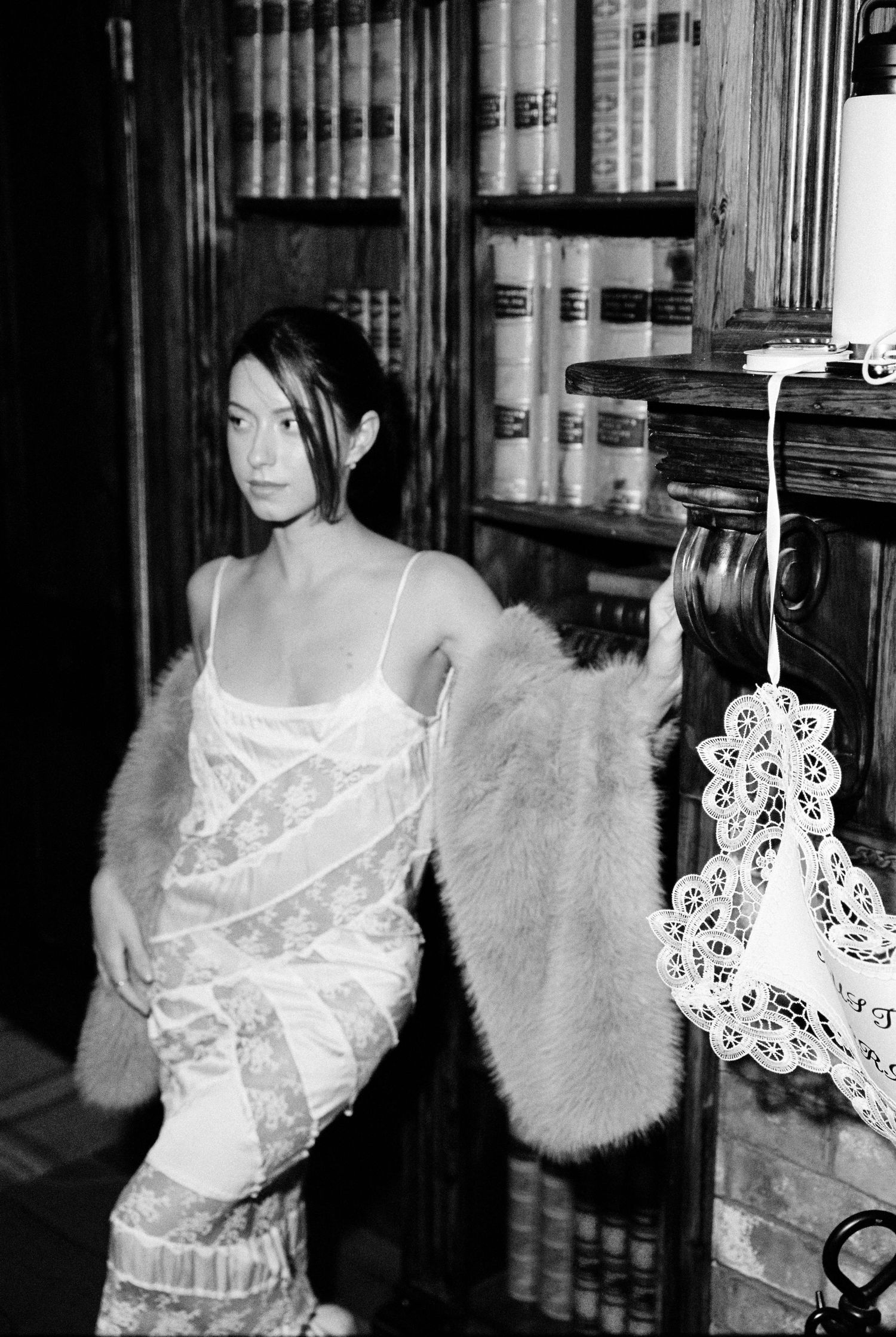 Elegant woman in slip dress with fur stole standing beside library bookshelf.