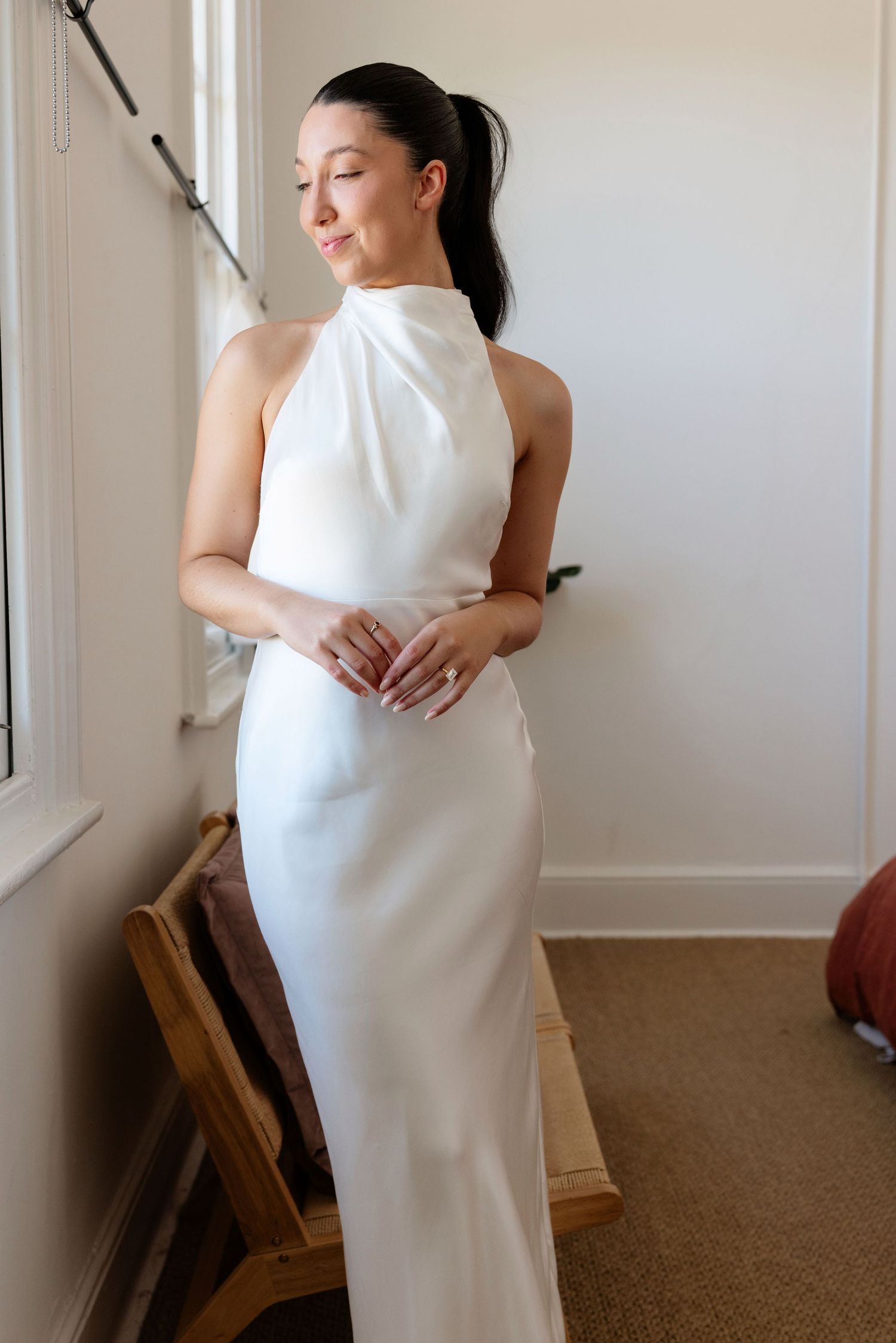 Bride in elegant white halter neck satin wedding gown standing by window with eyes downcast.