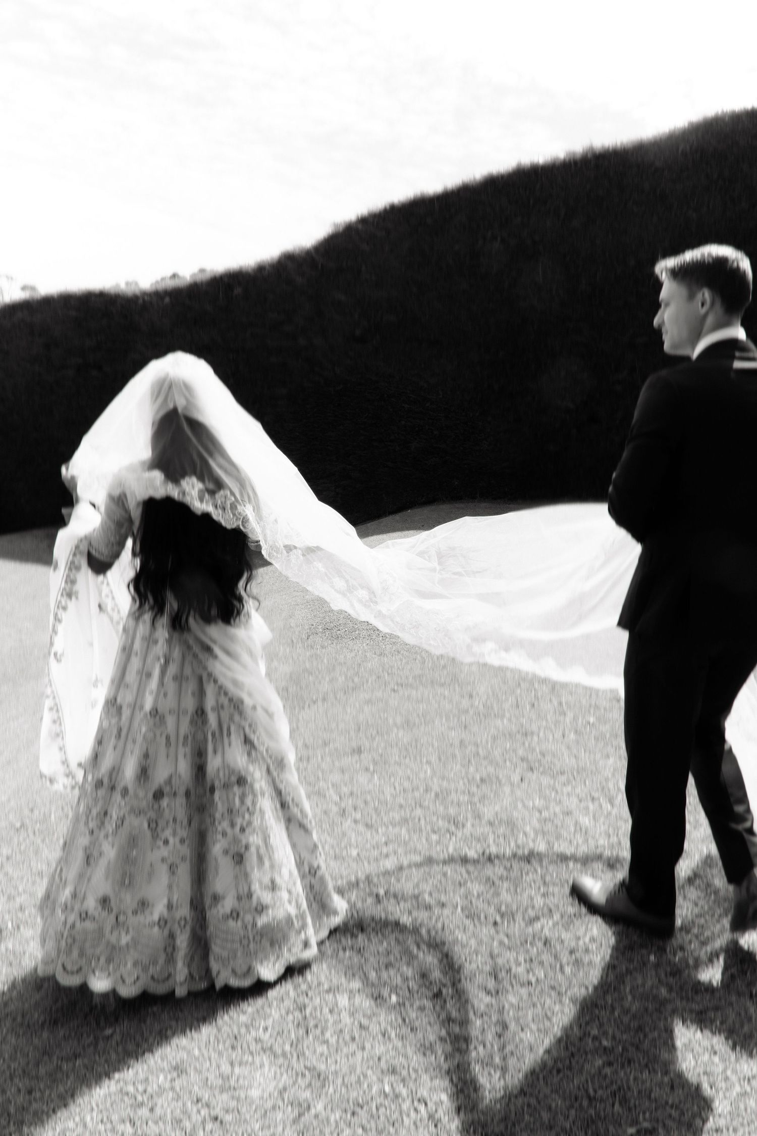 Black and white photo of bride in lace gown with flowing veil walking with groom outdoors.