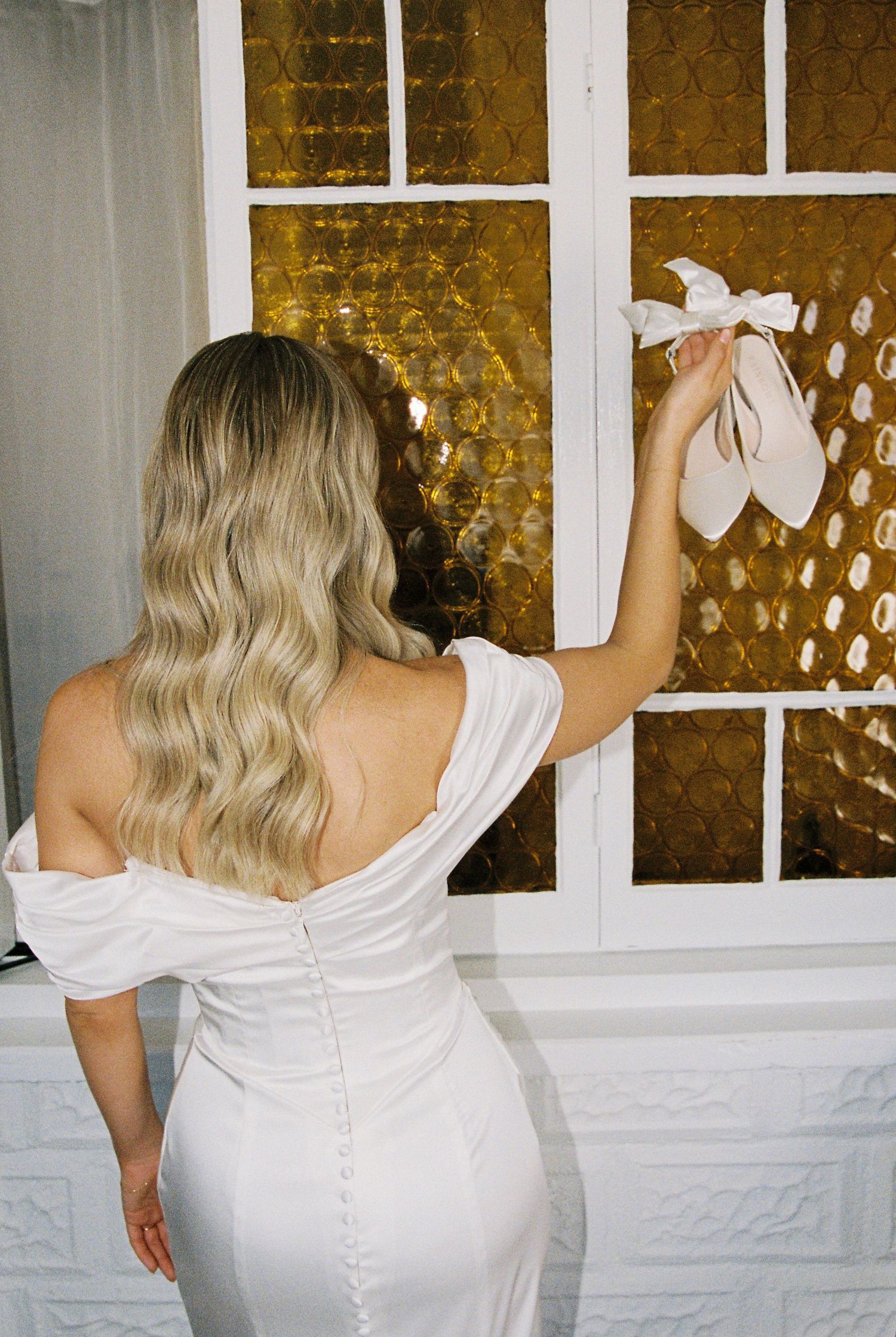 Bride in white off-shoulder gown holds bridal heels up to golden stained glass window.