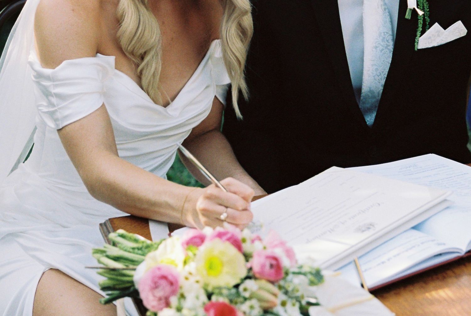 Bride in white off-shoulder dress signing wedding certificate with bouquet of pink flowers in foreground.