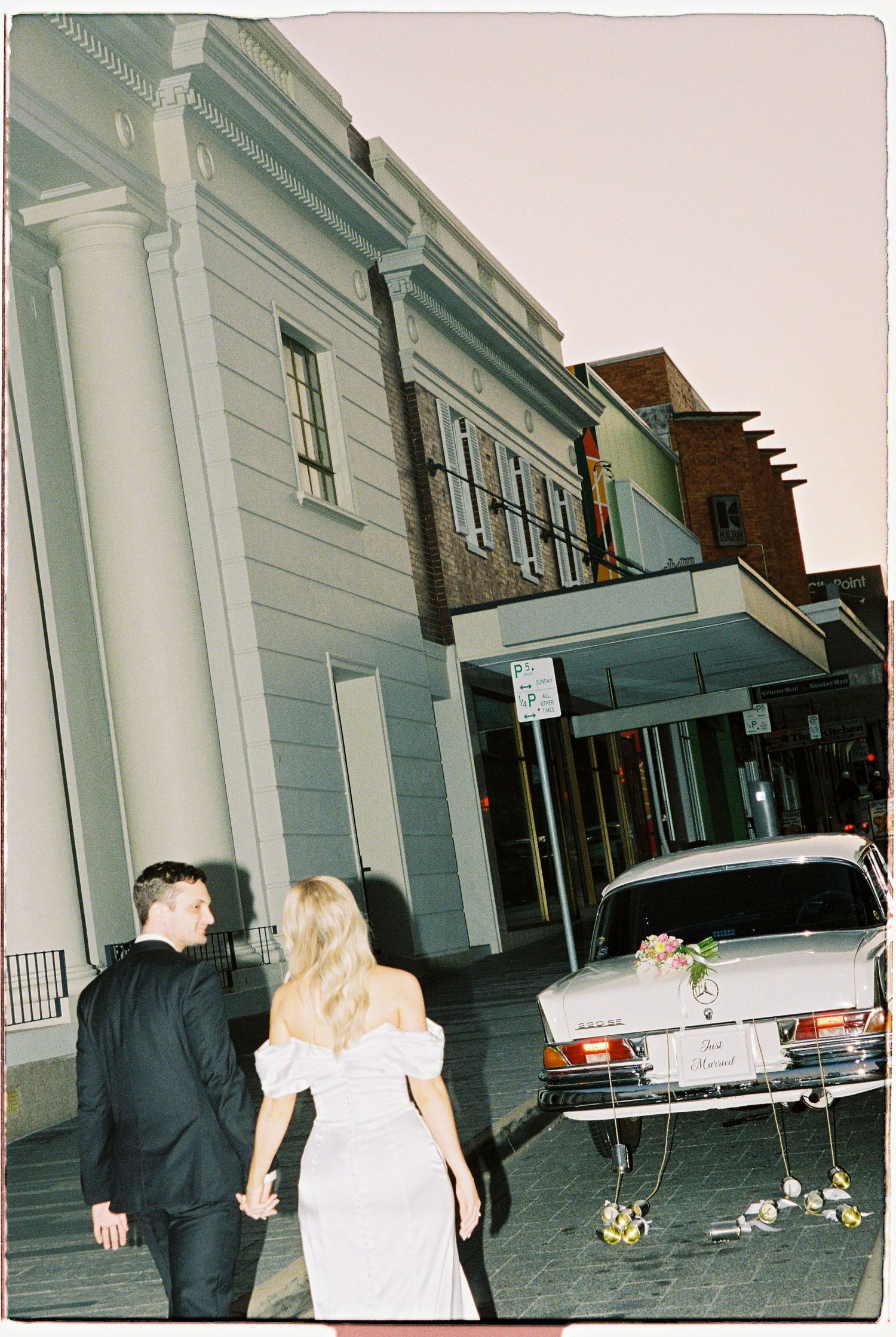 Bride and groom walking hand-in-hand toward venue beside vintage white Mercedes wedding car.