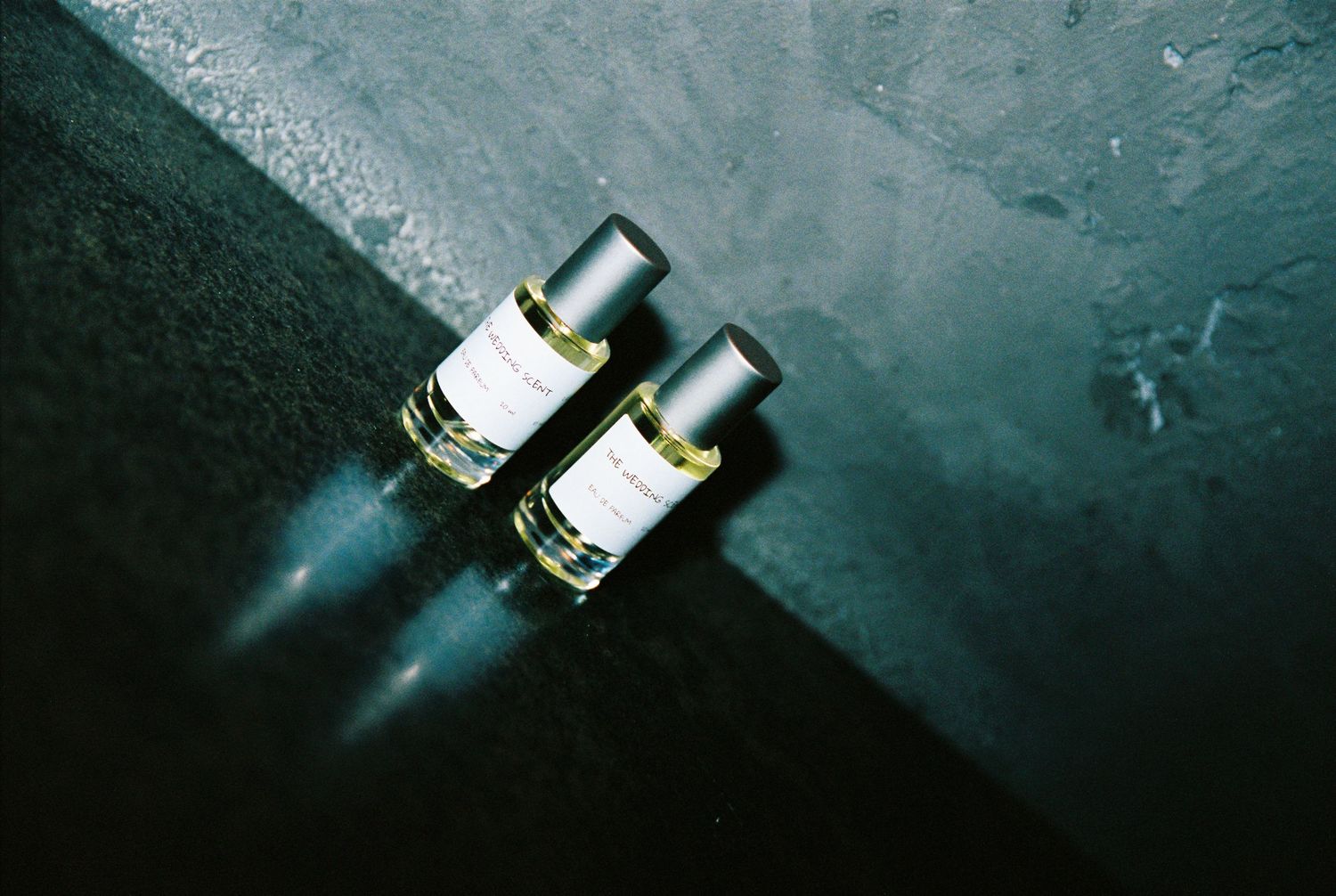 Two small perfume bottles casting shadows on a reflective dark teal surface.