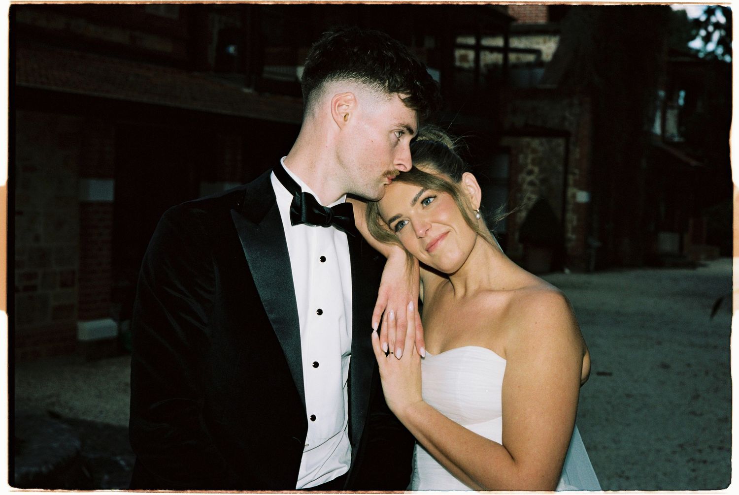 Romantic bride and groom in black tuxedo share intimate embrace at dusk on their wedding day.