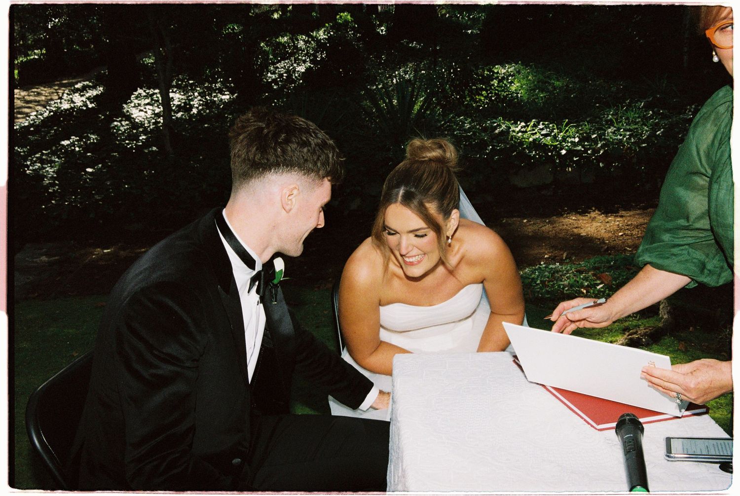 A bride in a white strapless gown and groom in a black tuxedo smile while signing their wedding certificate outdoors.