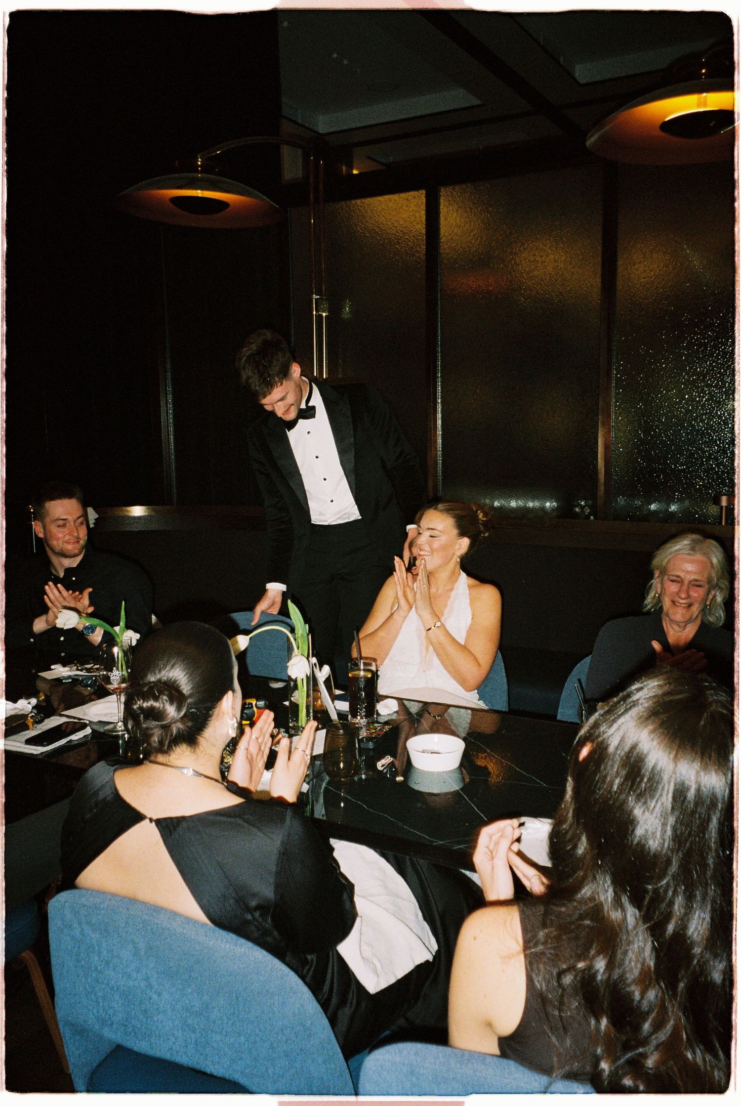 Groom in tuxedo presenting gift to emotional bride seated at elegant restaurant dinner table.