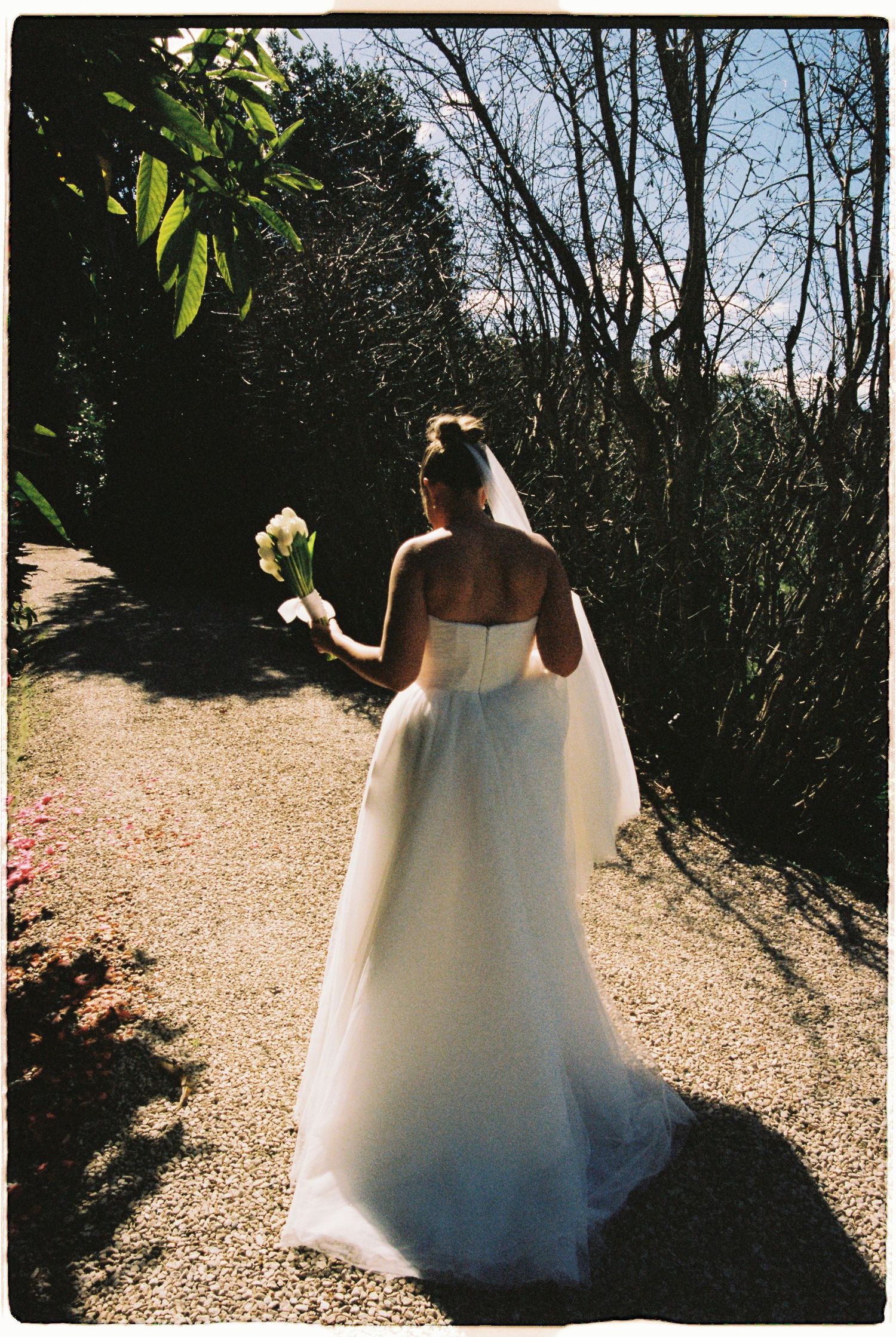 Bride in strapless white wedding gown walks along garden path holding a white floral bouquet.