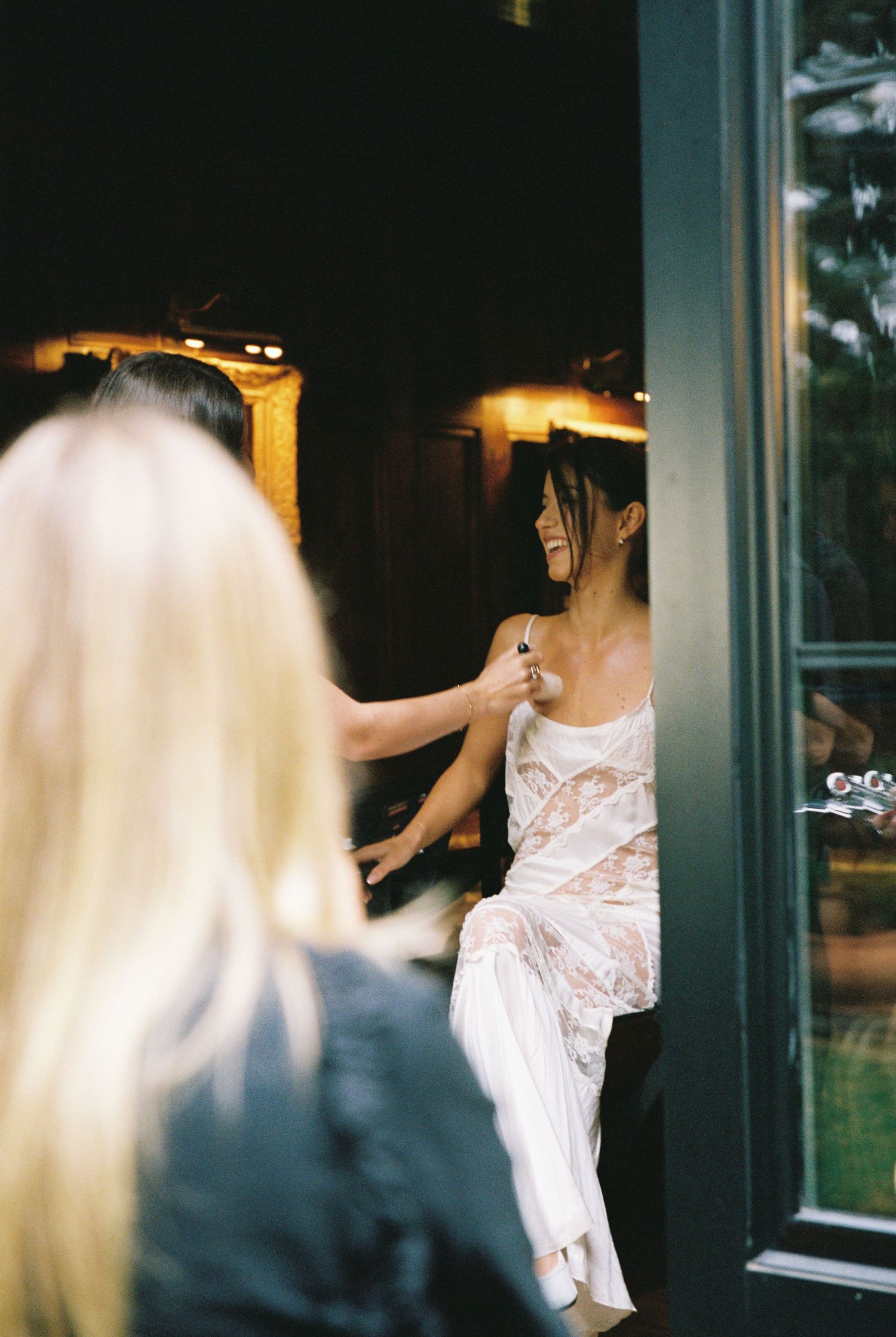Bride in white lace dress being helped by blonde bridesmaid in doorway before wedding.