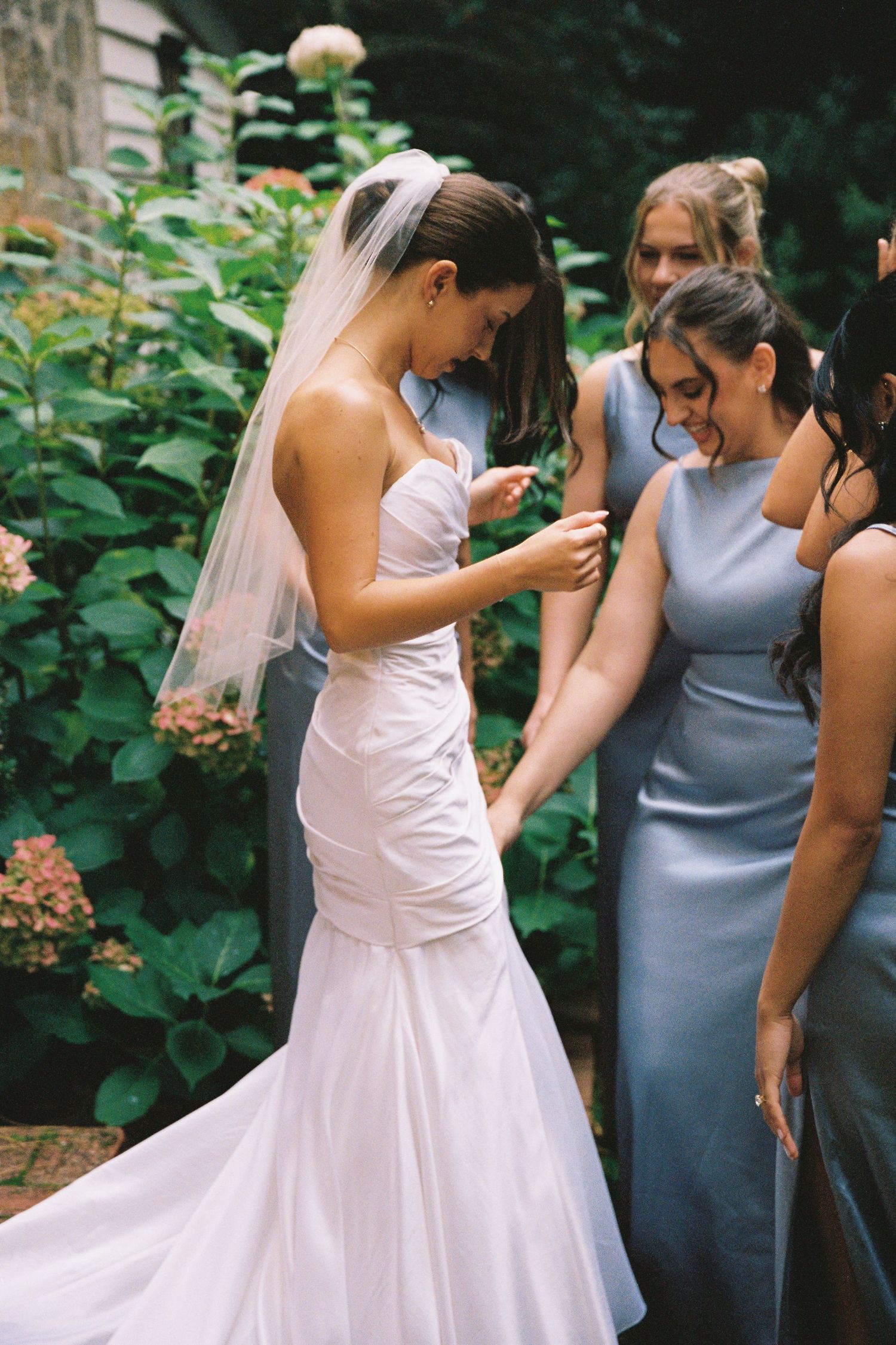 Bride in strapless mermaid wedding gown with veil assisted by bridesmaids in blue dresses outdoors.