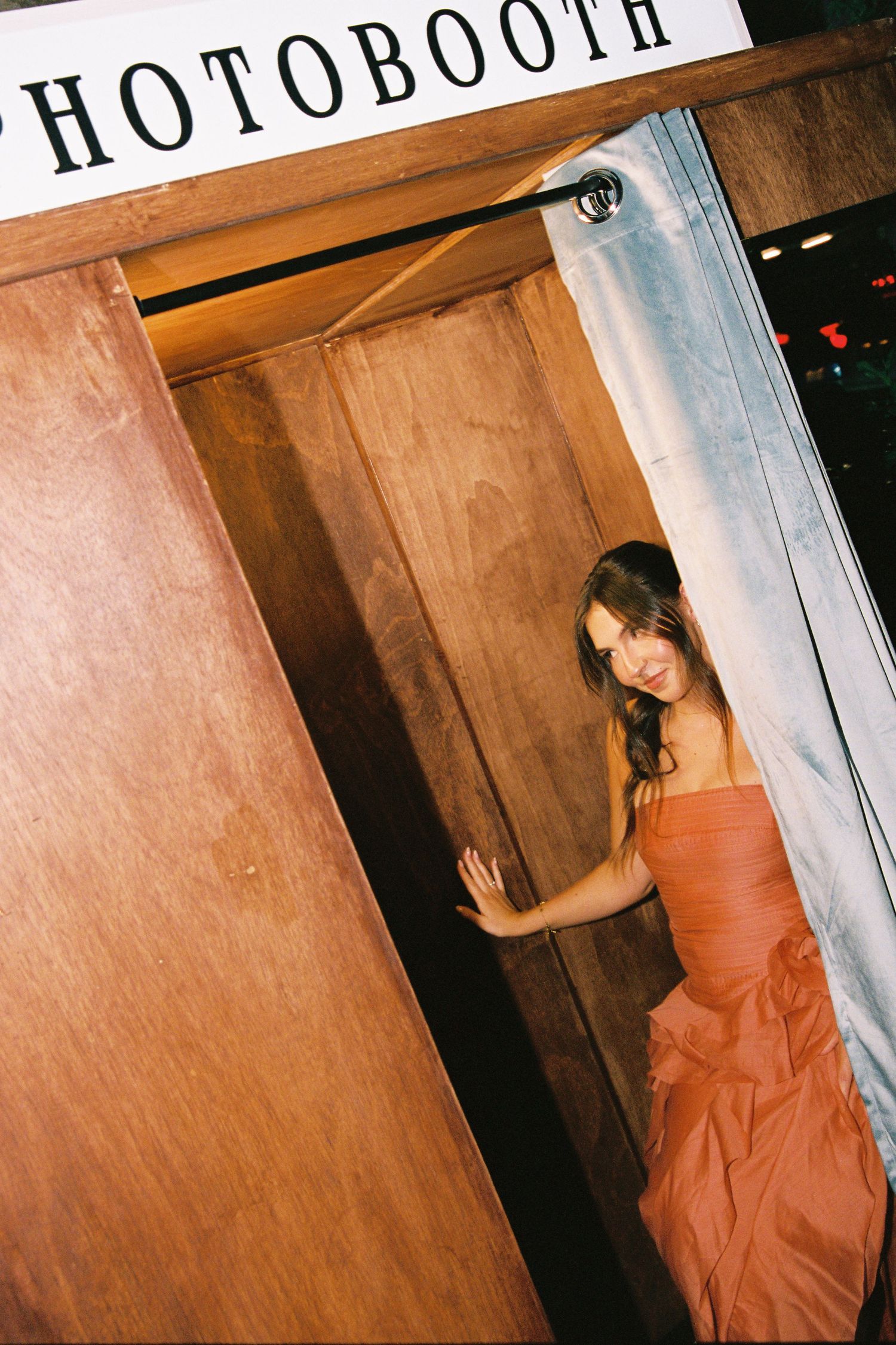 Woman in orange strapless gown posing at entrance of wooden photo booth curtain.